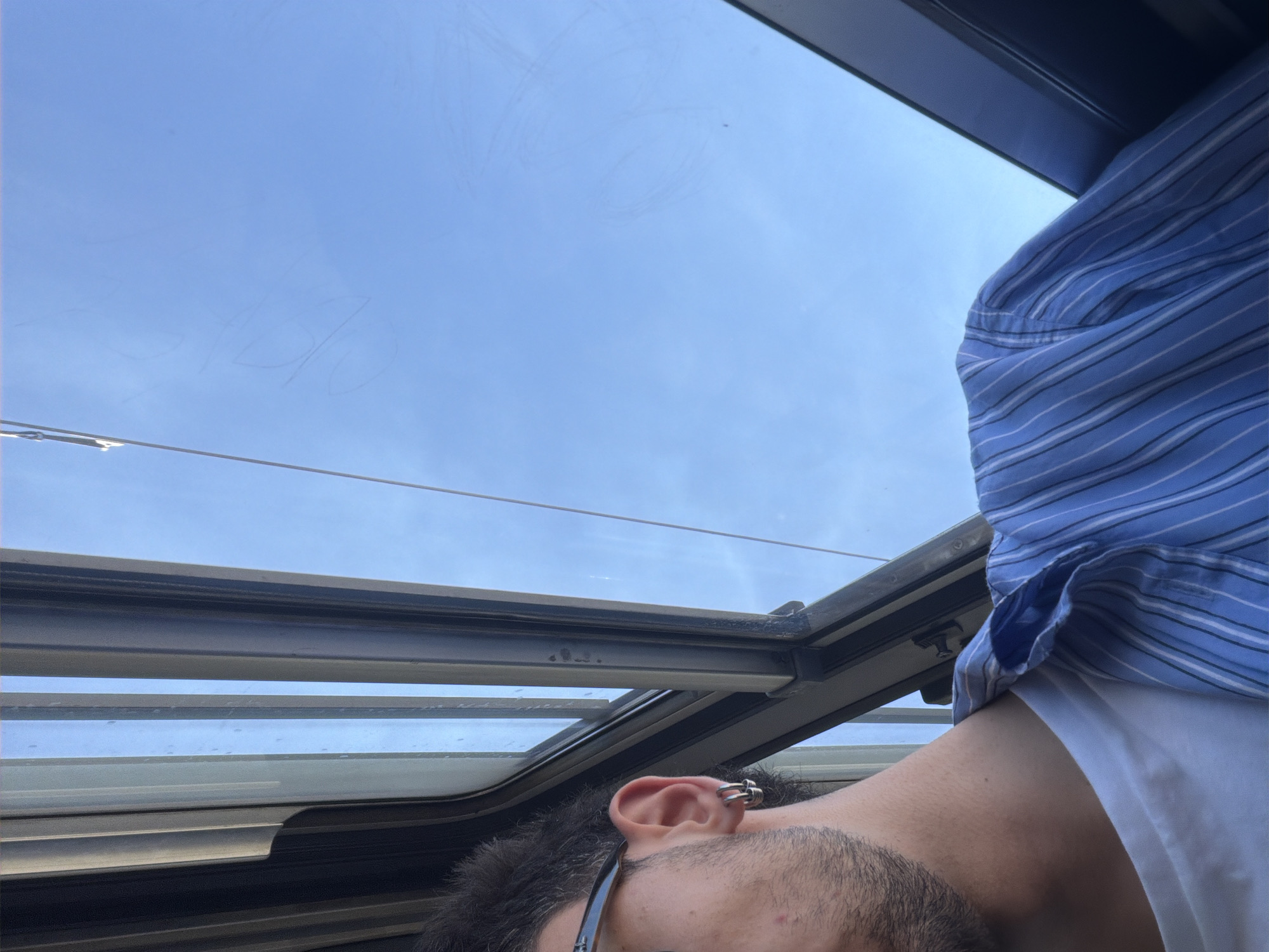 An individual is positioned inside a vehicle, looking upwards through a sunroof toward a blue sky. The person has dark hair and a fair complexion, and wears glasses and a blue striped shirt. The vehicle's sunroof is partially open, revealing the clear sky. The location is possibly Milan, Italy, although no specific features confirm this.