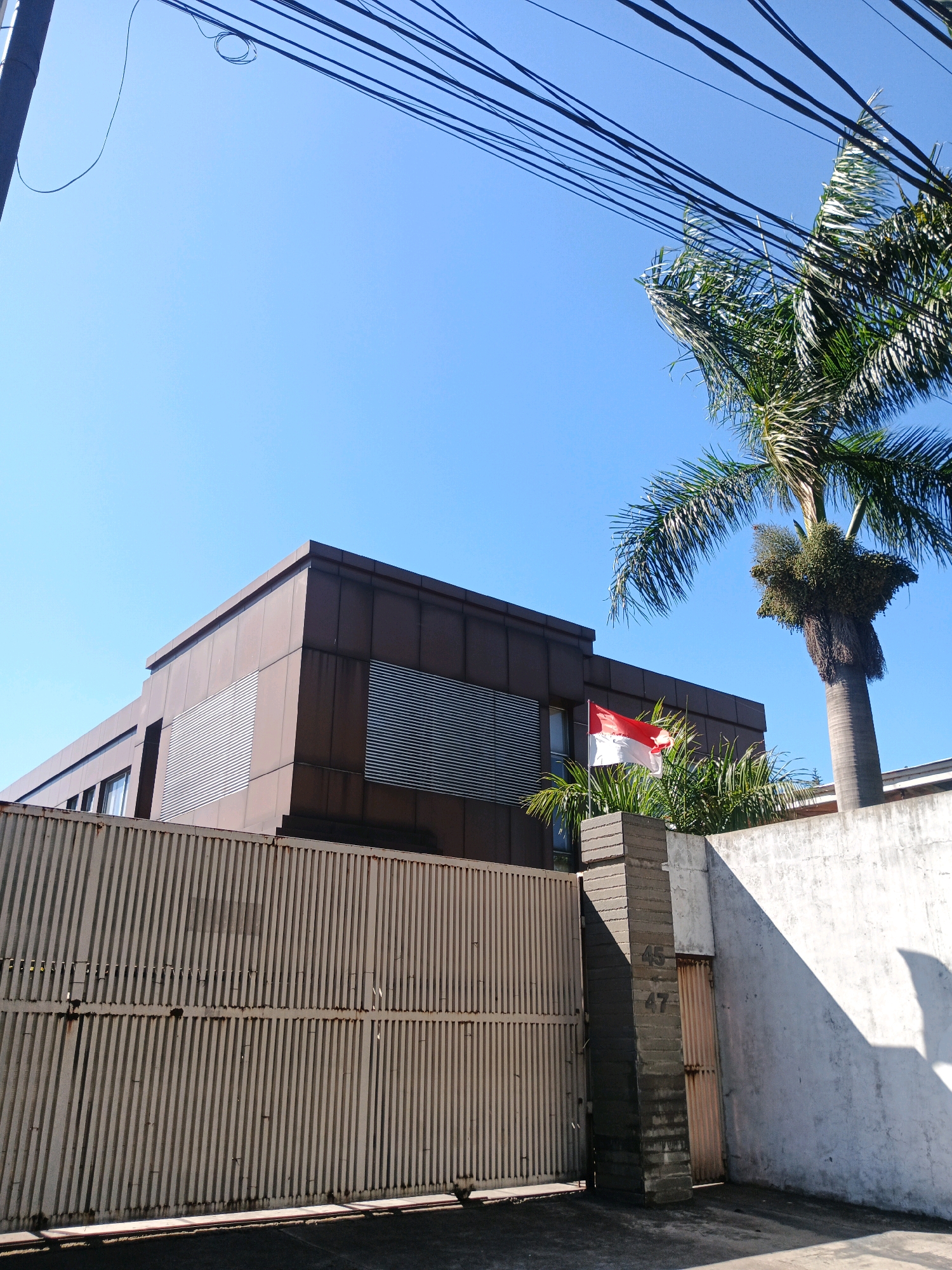 The image depicts a brown, modern building with horizontal louvers on its facade, seen from the exterior. A tall metal gate with vertical slats runs across the foreground. To the right of the gate, a concrete pillar displays the numbers "45" and "47". A palm tree stands beside the building, and an Indonesian flag (red and white) is visible on a flagpole near the gate. The sky is clear and blue, suggesting a sunny day. The setting appears to be a residential or commercial property within a city, possibly Bandung City, Indonesia, given the Indonesian flag. Overhead, a cluster of power lines is visible against the sky. The scene is static and captured in daylight.