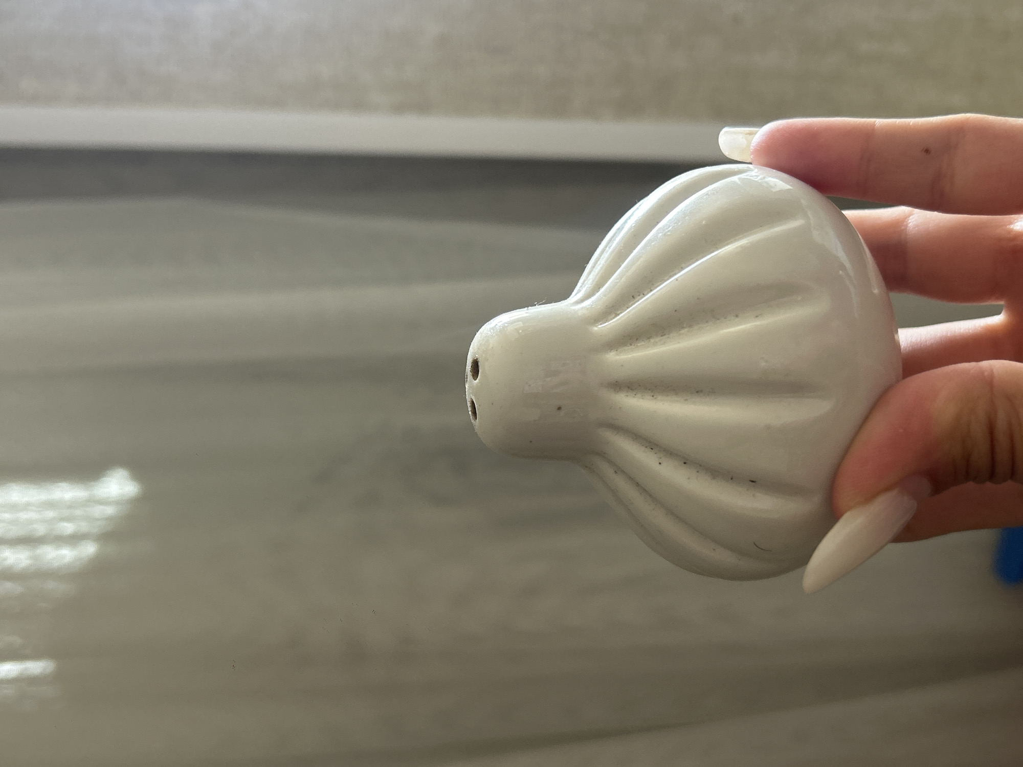 A white ceramic salt shaker, shaped like a traditional Khinkali dumpling, is held by a person's hand. The salt shaker has vertical ridges and a narrow top with three small holes. It is partially obscured by the thumb and index finger of the hand, which has long, light-colored acrylic nails. The background consists of a light-colored, sheer curtain, with vertical folds and bright light filtering through it, suggesting an indoor setting, possibly a home or studio apartment in Khyrdalan, Azerbaijan. The lighting creates a soft, diffused effect.