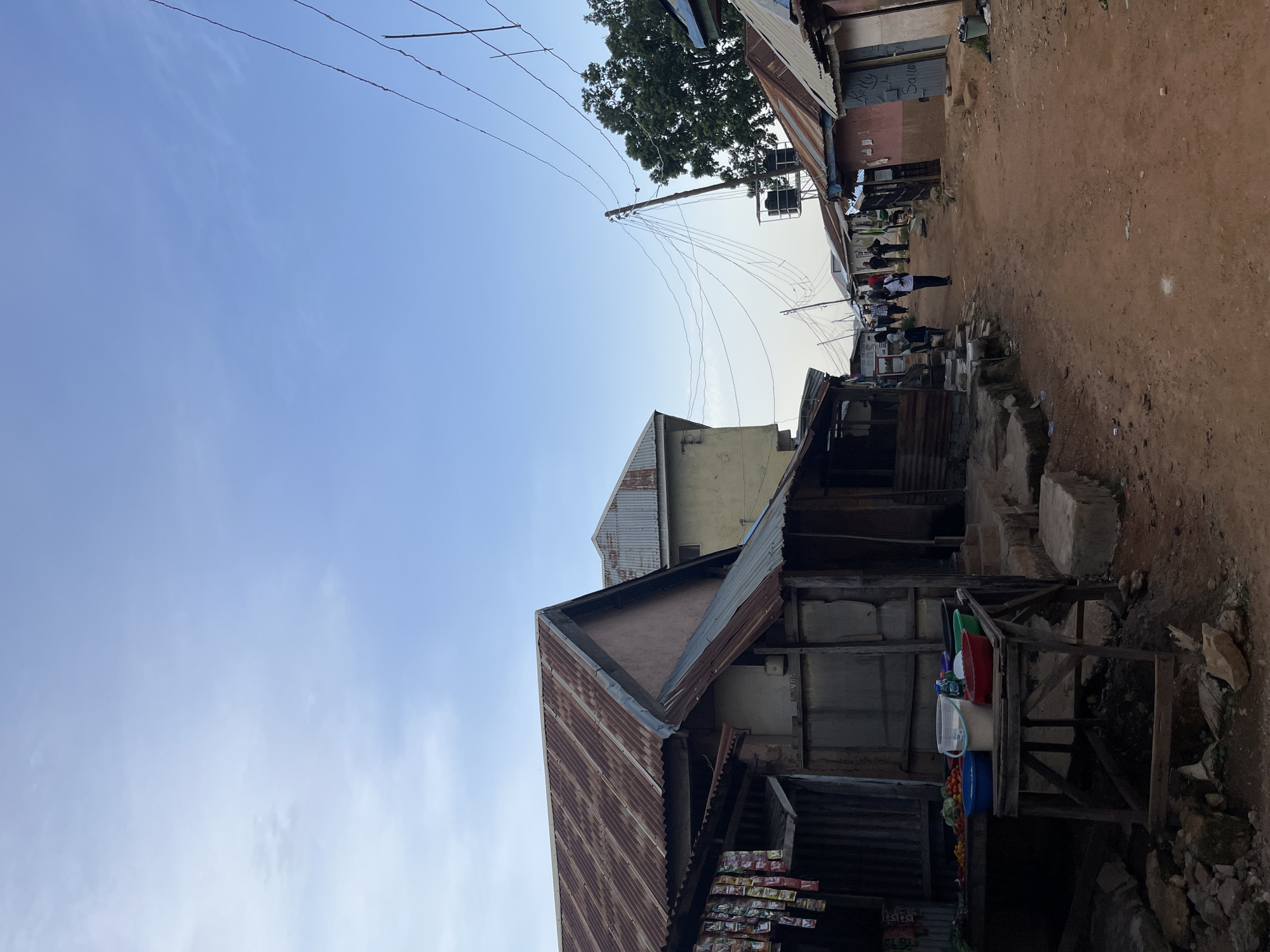 The image shows a street scene in Jos, Nigeria, under a clear blue sky. The perspective is from a low angle, looking up and slightly to the right. A dirt road runs through the middle of the scene, lined with makeshift buildings constructed from corrugated metal and other scavenged materials.
Several people are walking down the road, some in small groups. They appear to be casually moving along the street, suggesting it is a normal day. The buildings on either side of the road have corrugated metal roofs, some of which appear rusted. One building in the foreground has a display of items, possibly goods for sale, at its base, including what looks like fruits and vegetables in a blue container.
Above the buildings, a jumble of electrical wires stretches across the sky, attached to a utility pole. A large tree with green foliage is visible in the upper left quadrant of the image, providing a stark contrast to the man-made structures. The overall impression is of a busy, everyday street in an area with developing infrastructure. The time of day appears to be morning or afternoon, given the brightness of the sky and the shadows. There is no visible text that can be clearly read.