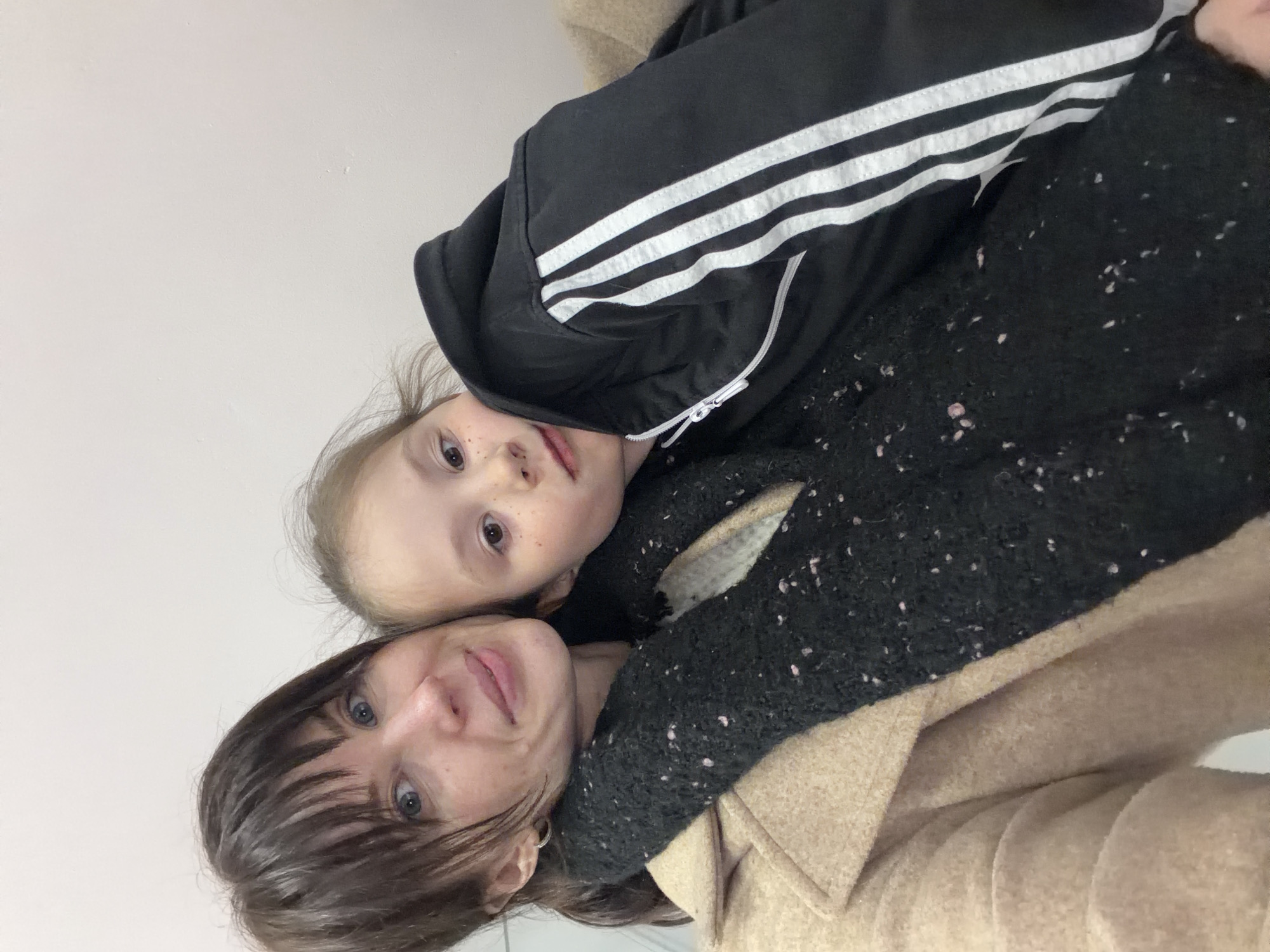 The image features two individuals, an adult woman and a child, captured in what appears to be a close-up selfie, presented with a vertical orientation (rotated 90 degrees counter-clockwise from a typical upright view).
The adult, likely the photographer, is visible in the lower portion of the frame (or the left side if viewed upright). She has dark hair with bangs and light-colored eyes. She wears a beige overcoat and a black, textured sweater with small light flecks. A silver hoop earring is visible on her left ear. Her expression is gentle, with a slight smile.
The child, positioned above the adult (or the right side if viewed upright), appears to be leaning against her. She has lighter hair, noticeable freckles on her face, and brown eyes. She is dressed in a black hooded jacket or tracksuit featuring distinctive white stripes on the sleeves and a visible zipper, characteristic of Adidas apparel. The child also looks towards the camera with a subtle, pleasant expression.
The background is a plain, light-colored wall, indicating an indoor setting. The overall mood conveys affection and closeness between the two subjects. There is no visible text in the image. The location context is Perm, Russia.
