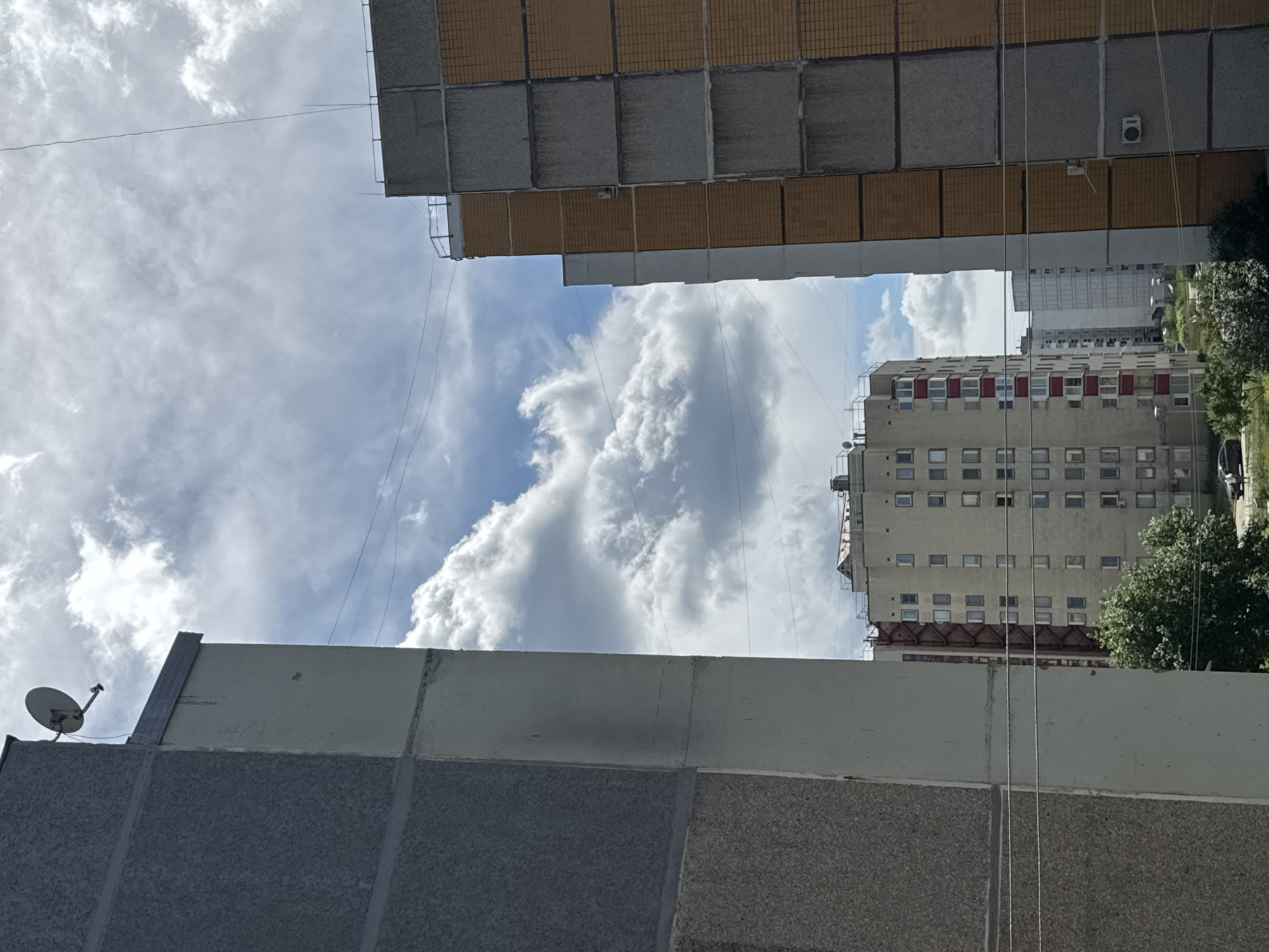 The image shows a view of apartment buildings in Chaikovsky, Russia. In the foreground, the left side of the frame is dominated by the textured gray facade of a building, with a lighter colored concrete vertical beam. On the top edge of this building, a satellite dish is visible. To the right, another building is partially visible, featuring a facade alternating between gray textured panels and orange brick-like panels. Further in the background, a light-colored apartment building with rows of windows and red accents is visible, along with other apartment buildings stretching into the distance. The sky is partly cloudy with a mix of blue sky and white cumulus clouds. Power lines crisscross the scene. At the bottom of the frame, some greenery from trees and a small section of a road with a parked car are visible.