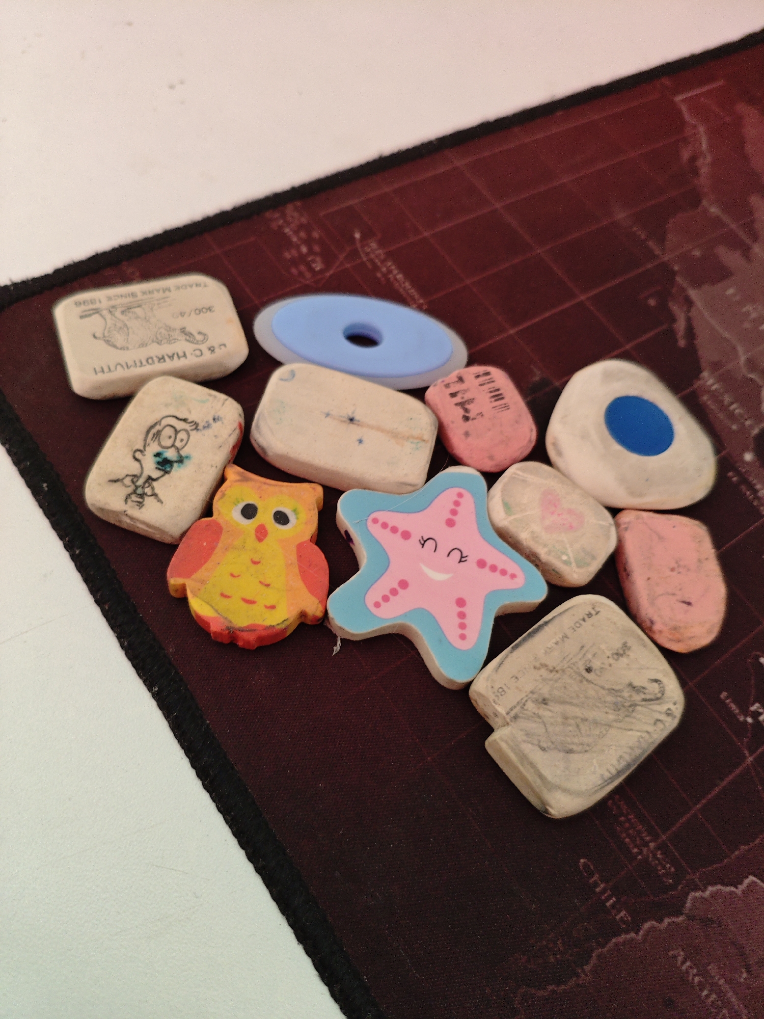 The image shows a collection of assorted erasers scattered on a dark red mousepad with a faded world map print. The erasers are of various shapes and colors, including a white rectangular one with writing and an image of an elephant, a blue disc-shaped eraser, a pink star-shaped eraser with a smiley face, an orange and yellow owl-shaped eraser, and several other plain and patterned erasers. The erasers are arranged in a scattered, slightly overlapping manner. The background is a white surface. The scene is indoors, likely on a desk. There are no people present in the image. The overall lighting is somewhat dim, casting slight shadows. The location in the city of Трудовское сельское поселение, Ukraine, is not depicted or suggested by any elements in the image.