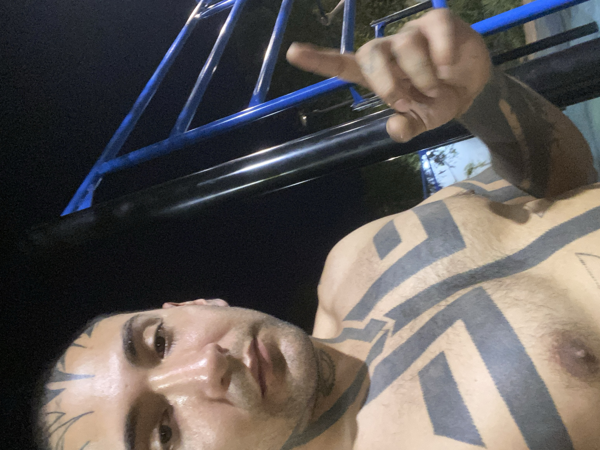 A close-up, low-angle selfie shows a shirtless man with extensive geometric tattoos on his chest, neck, and face. He is holding up his middle finger towards the camera. The background shows a metal structure with blue painted supports. The scene appears to be outdoors, with dark lighting. The exact location in Buenos Aires is not discernible.