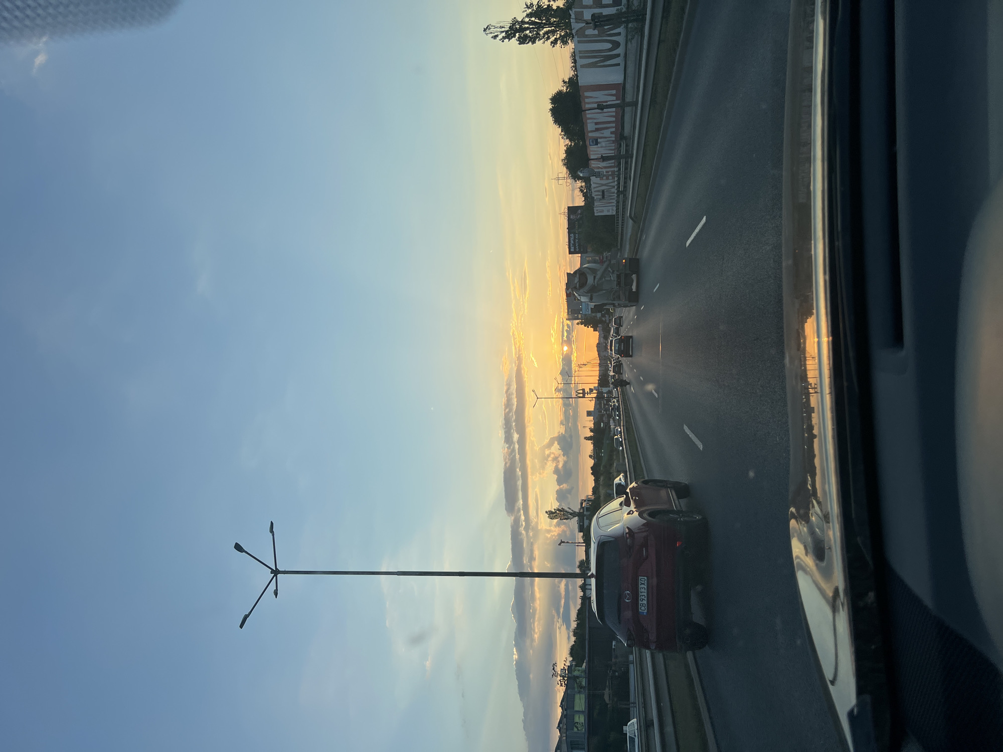 This media file depicts a sunset view from a vehicle on a highway in Sofia, Bulgaria. The sun is low on the horizon, casting a warm, golden light across the sky and illuminating the clouds with vibrant hues of orange and yellow. The scene is dominated by the highway, with several cars visible, suggesting moderate traffic. A red SUV is prominently featured in the foreground, its license plate visible with the characters "CB 9313 XO". A lamppost with three lights stands tall on the left side of the image. Billboards are visible along the side of the highway, one of which displays text that appears to be a mix of Latin and Cyrillic characters, reading "NUR" and some other Cyrillic words. The overall mood is serene, with the beauty of the sunset creating a peaceful atmosphere. The weather appears clear, with some scattered clouds adding to the visual appeal of the sky. The time of day is clearly sunset.