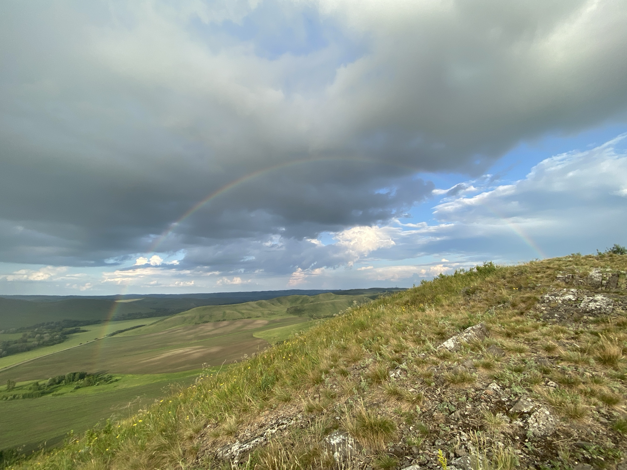 A wide-angle, slightly low-angle shot captures a vast, rolling landscape under a dramatic sky. In the foreground, the top of a rocky, grassy hill slopes down to the right, with dry, golden grasses and scattered rocks dominating the texture. A faint rainbow arc is visible in the sky above the distant green hills and fields, suggesting recent or ongoing rain. The sky itself is a mix of heavy, dark grey clouds on the left and brighter, lighter clouds with patches of blue sky breaking through on the right. The distant landscape consists of gently undulating hills covered in green fields and some patches of darker vegetation, possibly trees. The overall impression is one of natural beauty and a dynamic, changeable weather system. The location is specified as the city of Petrovskoe, Russia.