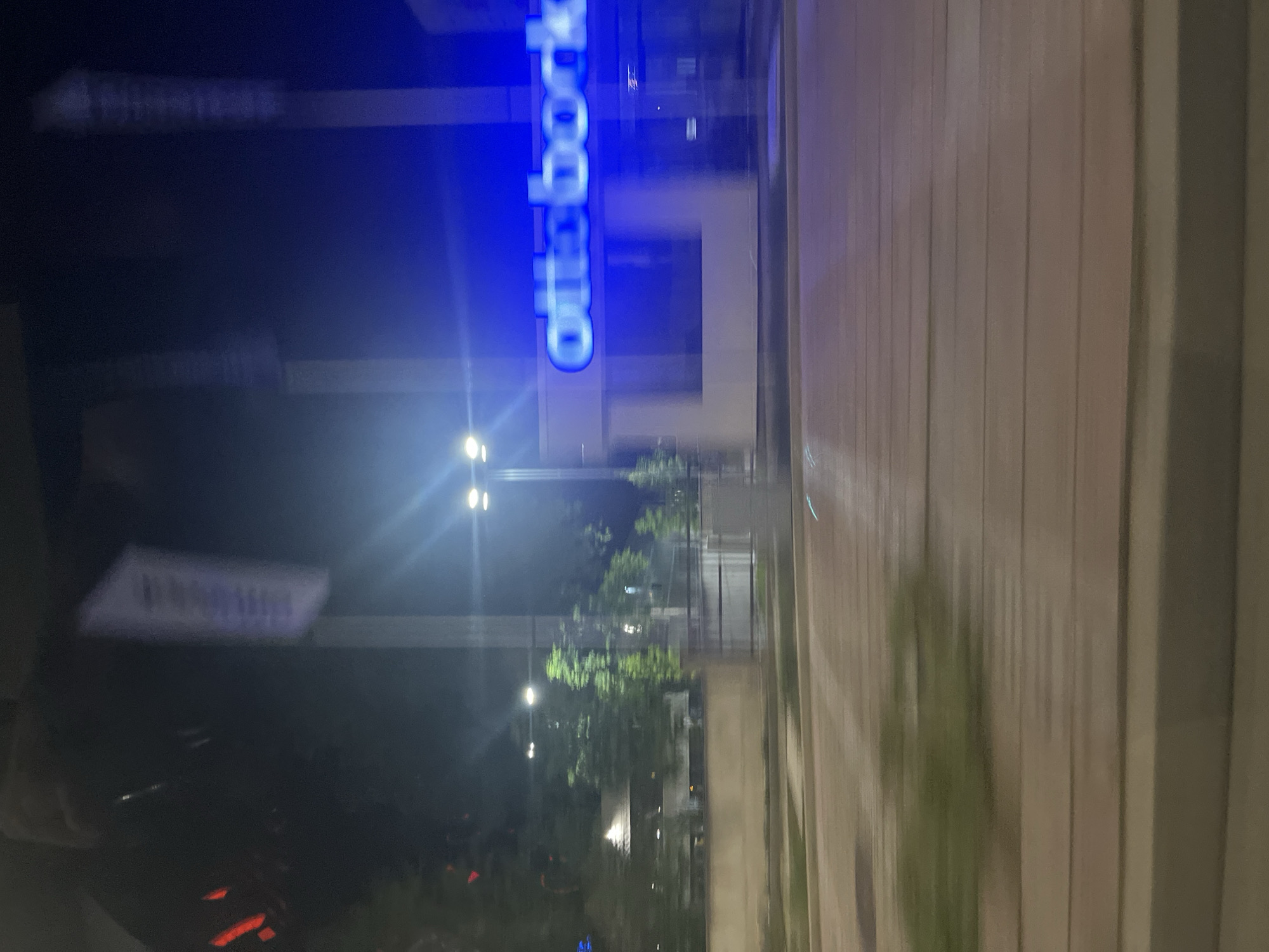 The image displays a nocturnal urban scene, captured with a vertical orientation, likely through a reflective surface such as a window, given the prominent reflections. The central focus is a vibrant blue neon sign, clearly legible as "ottobock." (the last character appears to be a full stop).
Behind the "ottobock" sign, the dark silhouette of a building is visible, with glimpses of lighter areas that could be windows or other architectural features. To the right of the sign, a light-colored, vertically-striped facade or wall dominates the right edge of the frame, alongside a paved surface, possibly a sidewalk or road.
In the middle and lower sections of the image, the dark environment is punctuated by several bright streetlights, which cast light onto sparse trees or bushes. These lights also create distinct lens flares. In the bottom-left corner, faint red lights are visible, suggesting the presence of a vehicle or another light source. Another blurry, rectangular white sign with unreadable text is also present in the lower left. The overall impression is an urban or commercial setting at night, illuminated by artificial light. The time of day is clearly nighttime.
