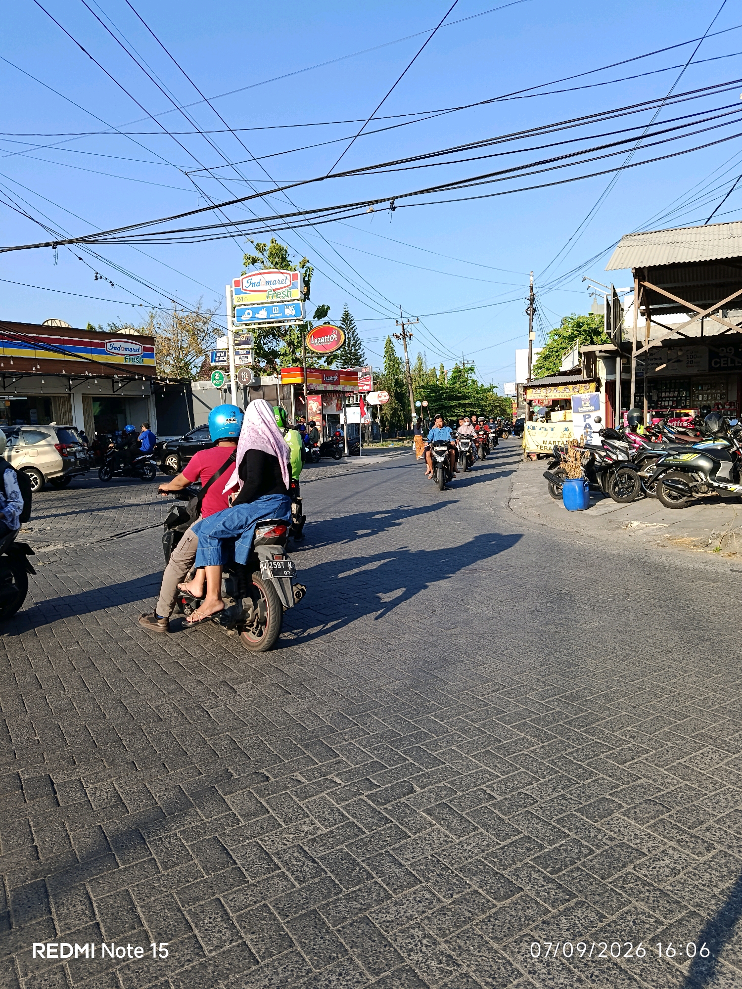 In Keboansikep, Indonesia, a street scene unfolds under a clear blue sky. A paved road, made of interlocking brick-like stones, fills the foreground and midground. The road is lined with shops and businesses on the right side, featuring awnings and various signs, including "Lazatto." On the left, a row of parked vehicles and storefronts, including an "Indomaret Fresh," are visible.
Several motorcycles are in motion. In the immediate foreground, a motorcycle carries two people: a man in a red shirt and blue helmet, and a woman in a light pink hijab and blue jeans. They are riding on the right side of the road, heading away from the viewer. Behind them, other motorcycles and their riders are visible further down the street. A blue plastic bin and some dried plant material are placed on the sidewalk to the right. The scene is illuminated by sunlight, casting shadows on the road. The time stamp "07/09/2026 16:06" is visible in the lower right corner. The text "REDMI Note 15" is in the lower left corner.