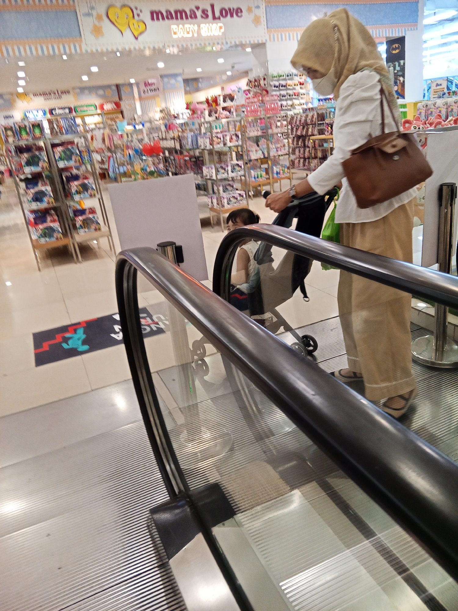 In an indoor shopping environment, likely a baby shop as indicated by the "mama's Love BABY SHOP" signage, a woman wearing a hijab, white shirt, and tan pants is pushing a stroller up an escalator. She has a face mask on and carries a brown purse over her shoulder. A small child is visible in the stroller. The escalator's metal handrail and glass panel are prominent in the foreground. The background shows shelves stocked with merchandise, including toys and baby products. The overall lighting suggests a well-lit commercial space. The presence of a "mama's Love BABY SHOP" suggests this location is likely in a mall or commercial center within Lubuklinggau, Indonesia.