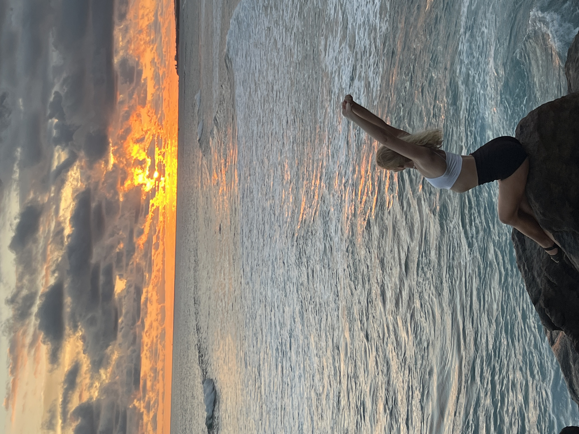 The image depicts a striking scene of a woman posing on a rock by the ocean at sunset. The woman, with blonde hair, is seen in profile, leaning forward with her arms extended upwards and slightly backwards, wearing a white crop top and black shorts. She appears to be in mid-motion, perhaps preparing to dive or performing a graceful stretch.
The background is dominated by a vibrant sunset, with the sun visible near the horizon, casting brilliant orange and yellow light across the sky and water. Dark grey clouds intermingle with the bright, warm-toned clouds, creating a dramatic sky. The ocean surface is rippled, reflecting the golden hues of the sunset. A faint line of sand is visible further along the distant coastline. The rock the woman is balancing on is dark and rugged.
The photograph captures a moment of natural beauty and human movement, set against the breathtaking backdrop of a tropical sunset. The location is contextualized as Goviyapana, Sri Lanka. There is no visible text in the media file.