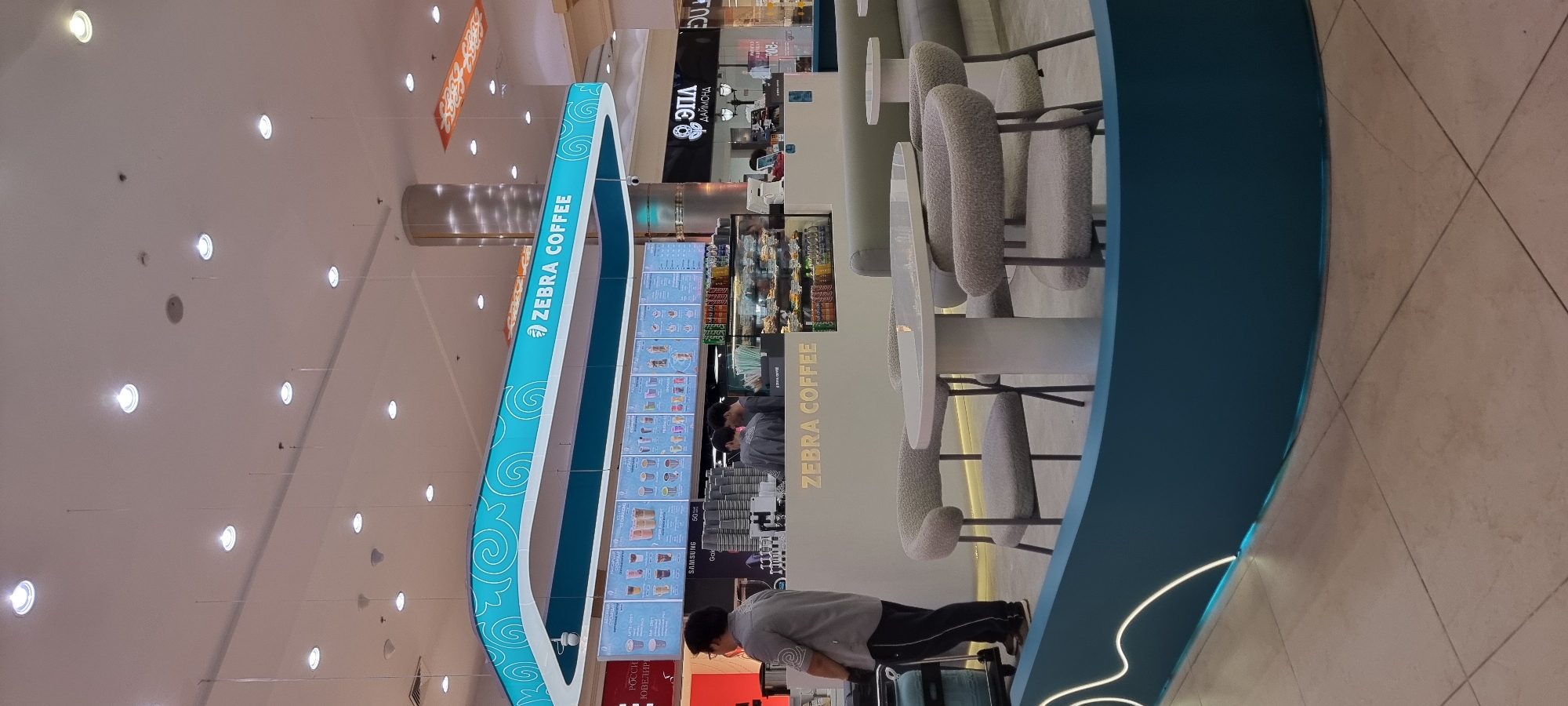 This image captures a view of a Zebra Coffee shop, likely located in the city of Aktobe, Kazakhstan. The scene is set indoors, possibly within a shopping mall, as suggested by the bright, overhead lighting and the general retail environment.
In the foreground, a modern, curved teal-colored barrier with integrated white lighting outlines a seating area. Two plush, grey armchairs are visible, with a contemporary white oval table between them, also illuminated from below by the barrier's lighting. The floor is tiled in a neutral beige color.
Behind this seating area, the Zebra Coffee counter is prominent. The counter itself is white, with the "ZEBRA COFFEE" logo displayed in gold lettering. Behind the counter, staff members are visible, appearing to be engaged in serving customers. Display cases filled with food items like pastries and drinks are also present.
Above the counter, a large, illuminated menu board displays various drinks and food options. The "ZEBRA COFFEE" logo is repeated on this signage. The overall aesthetic is clean, modern, and inviting, with a focus on bright lighting and contemporary design elements. The presence of other retail establishments and signage in the background further reinforces the idea of an indoor shopping or commercial complex.