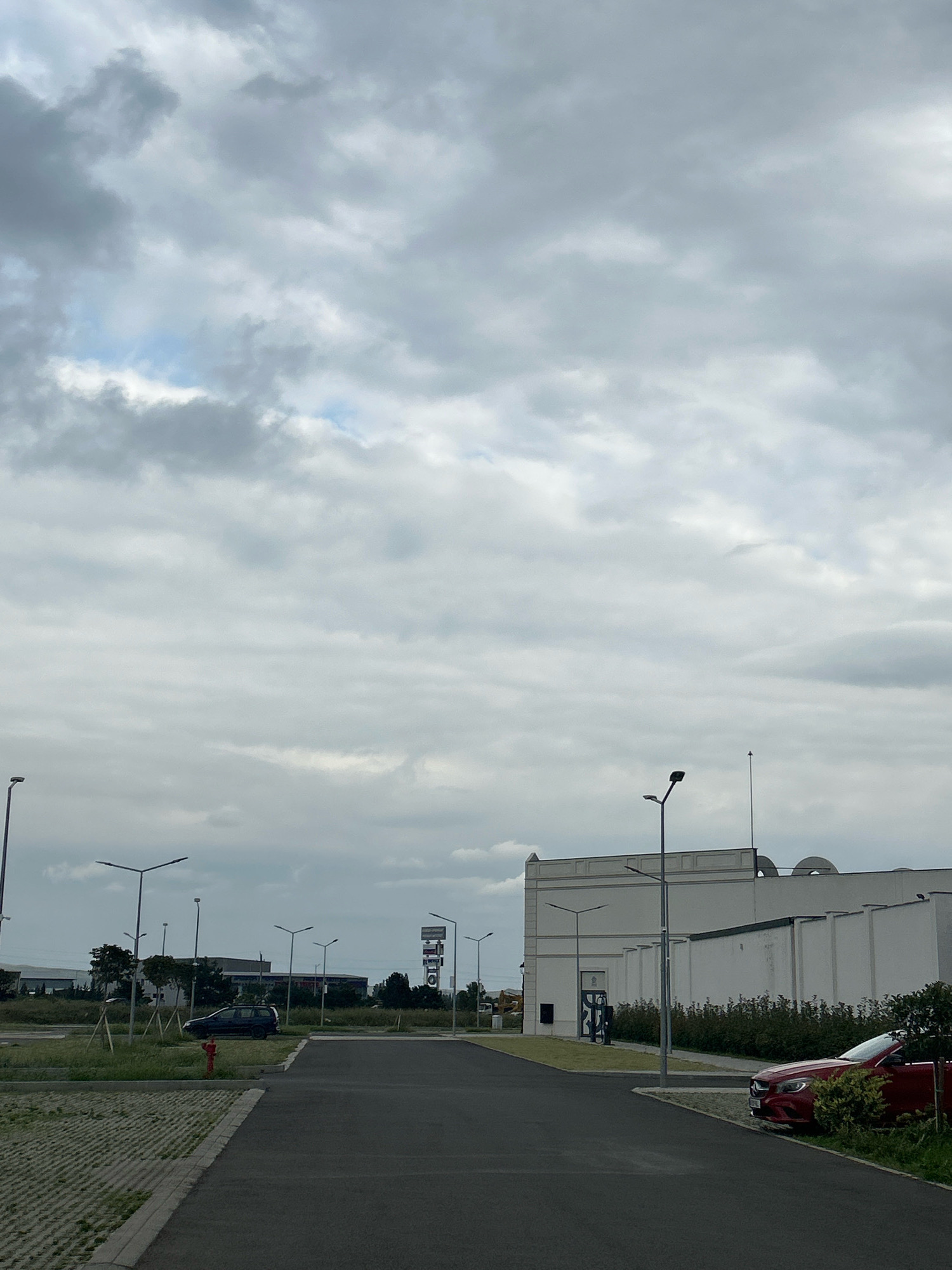 The image depicts an outdoor scene under a cloudy sky, likely in the city of Gamarjveba, Georgia, given the context. In the foreground, there is a paved road leading towards the right. To the left of the road is a parking area with a permeable paving surface. A dark-colored SUV is parked on the left side of the road, near a red fire hydrant and some grass. Further to the left, there are numerous streetlights and some low-lying buildings and trees.
To the right of the road, a modern, white building with a flat roof and some decorative elements on its facade is prominent. There are also several streetlights in front of this building. A red convertible is parked on the right edge of the paved area, partially obscured by bushes. The background consists of more buildings and vegetation, as well as more streetlights and a signpost. The overall impression is that of a commercial or industrial area, possibly a retail park or business complex.