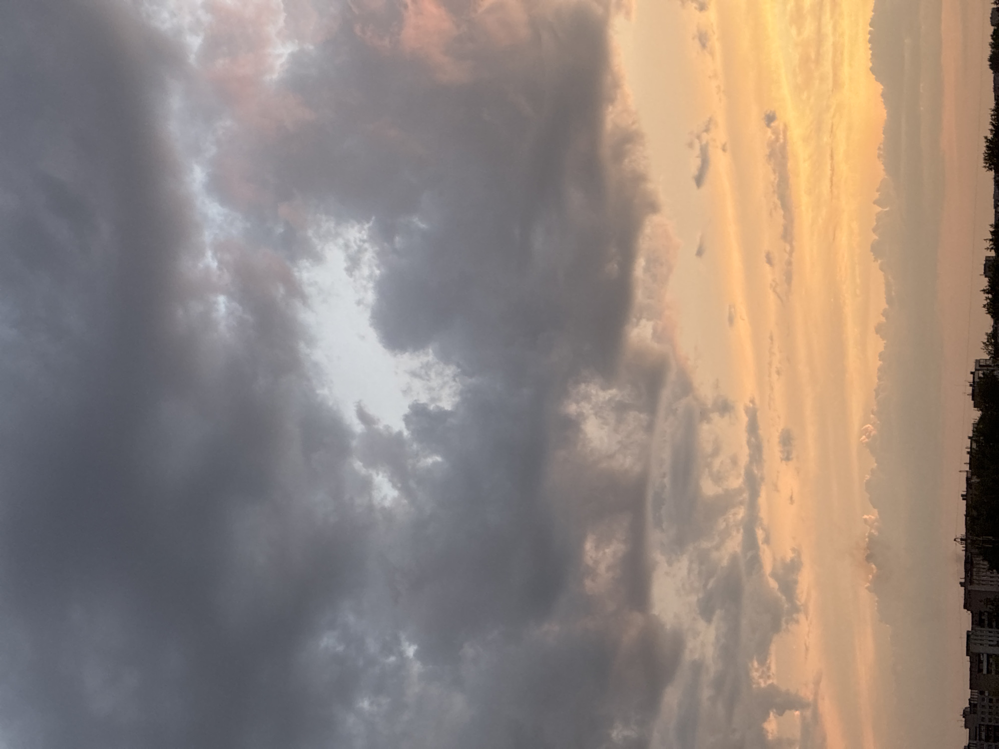 The image shows a cloudy sky above a city skyline. The sky is filled with a mix of dark gray and light gray clouds, with some patches of pink and orange light from the setting sun peeking through. The clouds are textured and appear to be moving. Below the sky, a silhouetted city skyline is visible, with buildings of varying heights and trees. The image is taken from a high vantage point, looking up at the sky. There are no people or animals visible in the image.