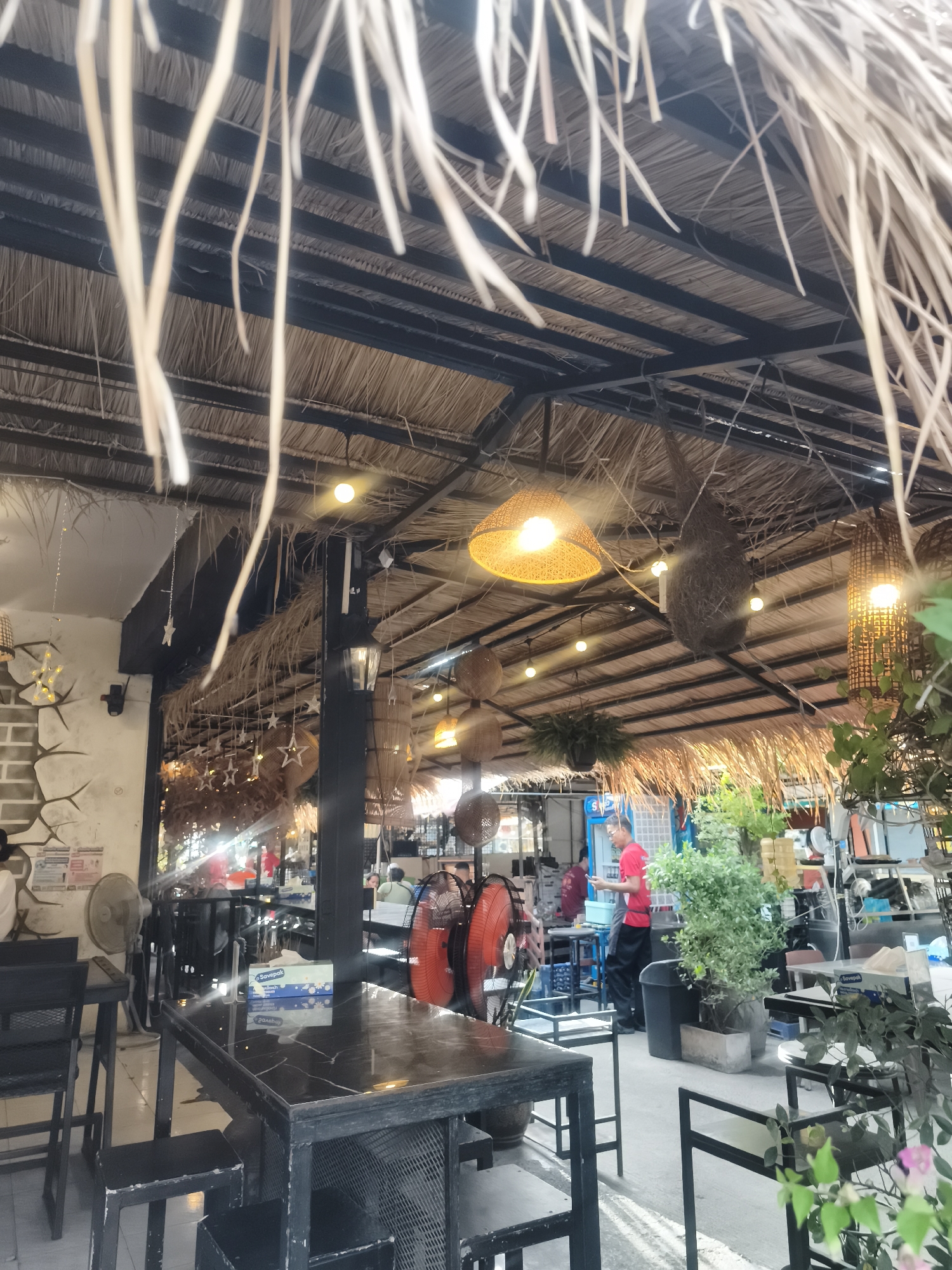 The image depicts an open-air restaurant in Baan Chaweng, Thailand. The roof is constructed with straw-like material over a black metal frame, with decorative straw strands hanging down. Woven lamps hang from the ceiling. The restaurant features black metal tables and chairs. A box of tissues sits on a table. People are present in the background; a man wearing a red shirt is standing near the back of the restaurant. Various plants in pots are scattered around the space. One fan is set on the ground.