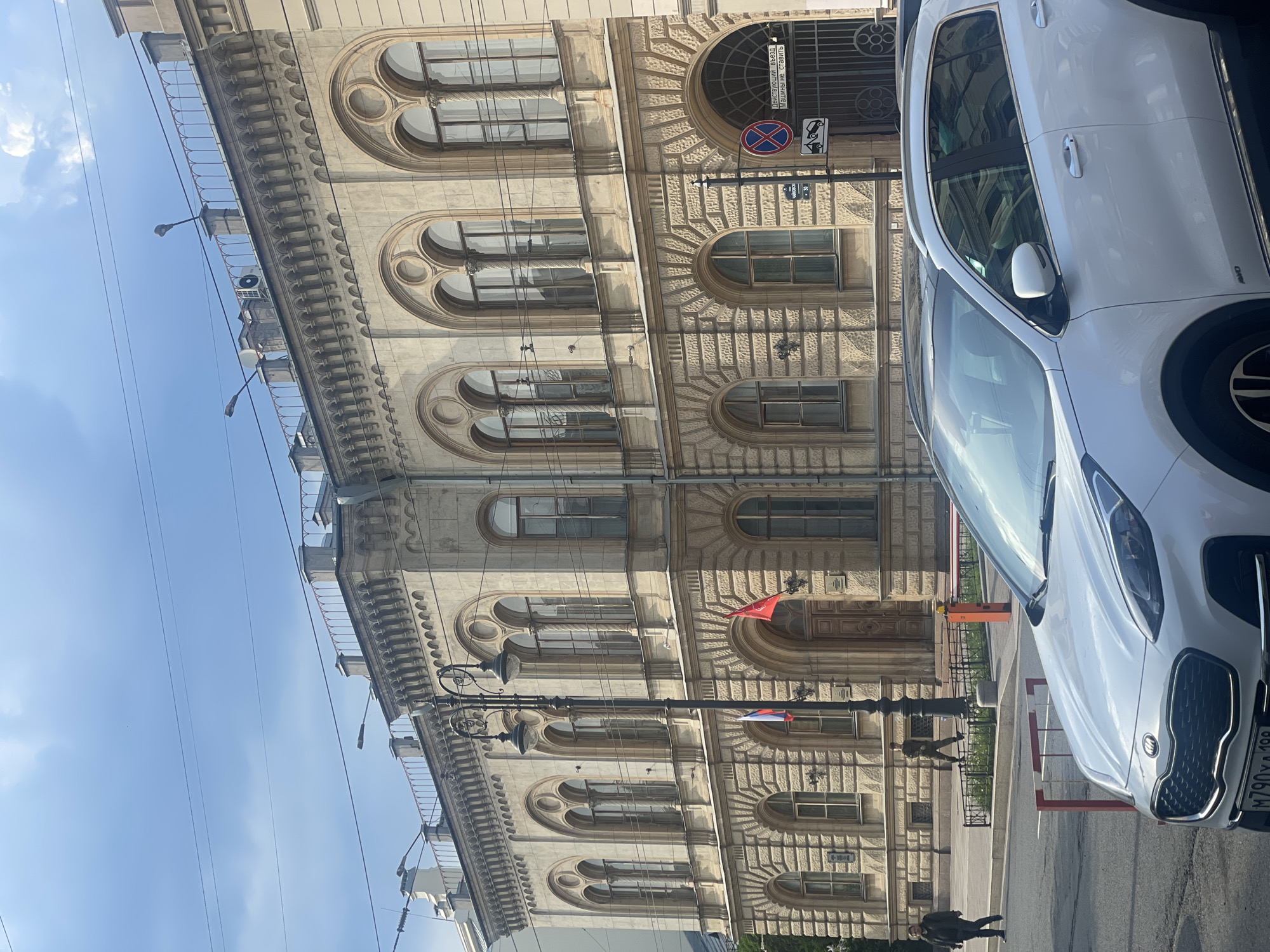 The image shows a building in Saint Petersburg, Russia. The building is light-colored with many windows in arched shapes. The architecture style appears to be historic. The street includes a white vehicle parked on the side of the street. There is a street lamp visible near the building with two small flags attached. One is red, and the other has three horizontal stripes. The sky is blue with some clouds. A no parking sign is also visible attached to the building.