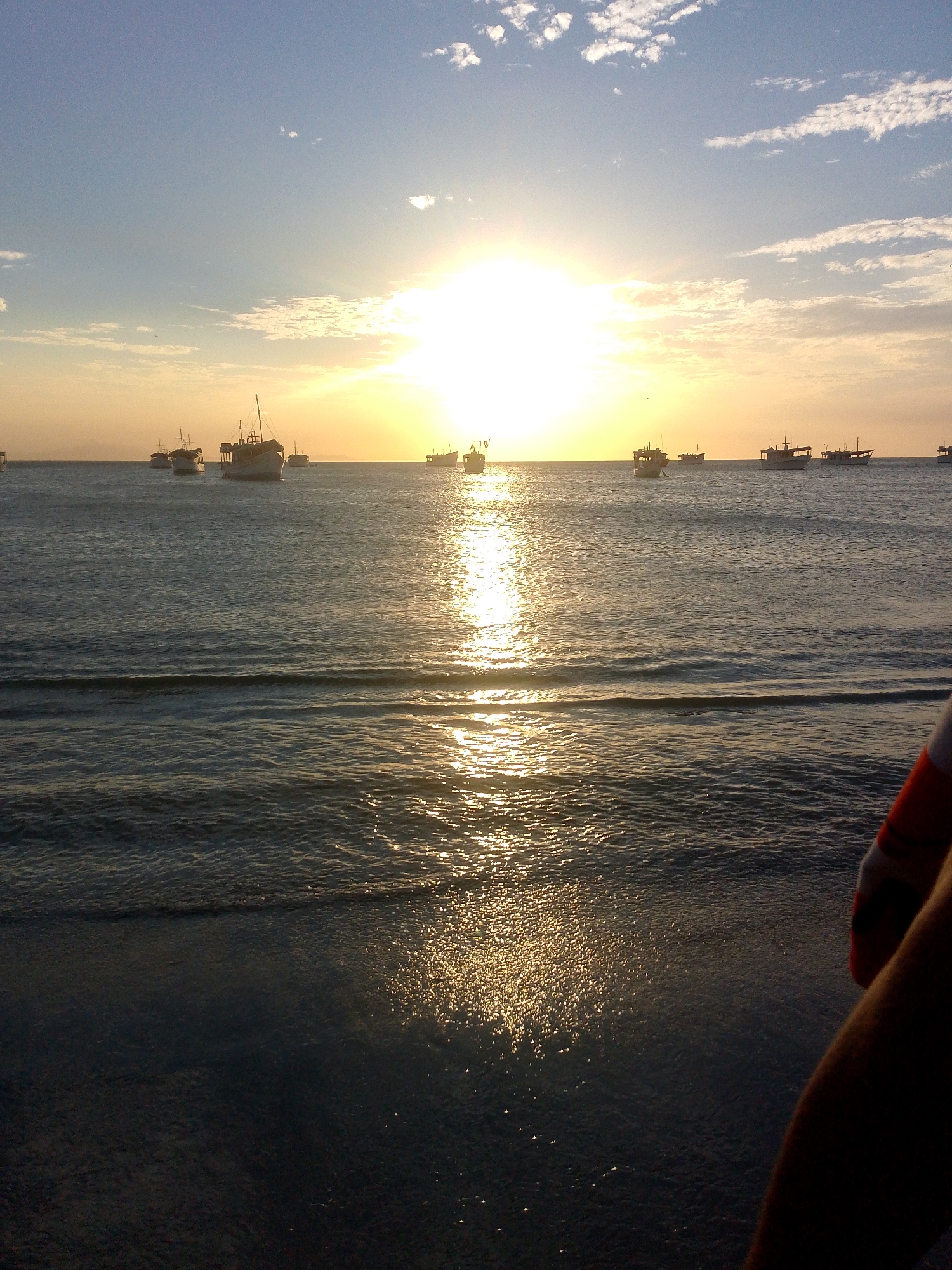 Captured in Juan Griego, Venezuela, the image showcases a sunset over the ocean, with multiple boats dotting the horizon. The sun's reflection creates a shimmering path on the water's surface. In the lower right corner, part of a person's arm is visible.