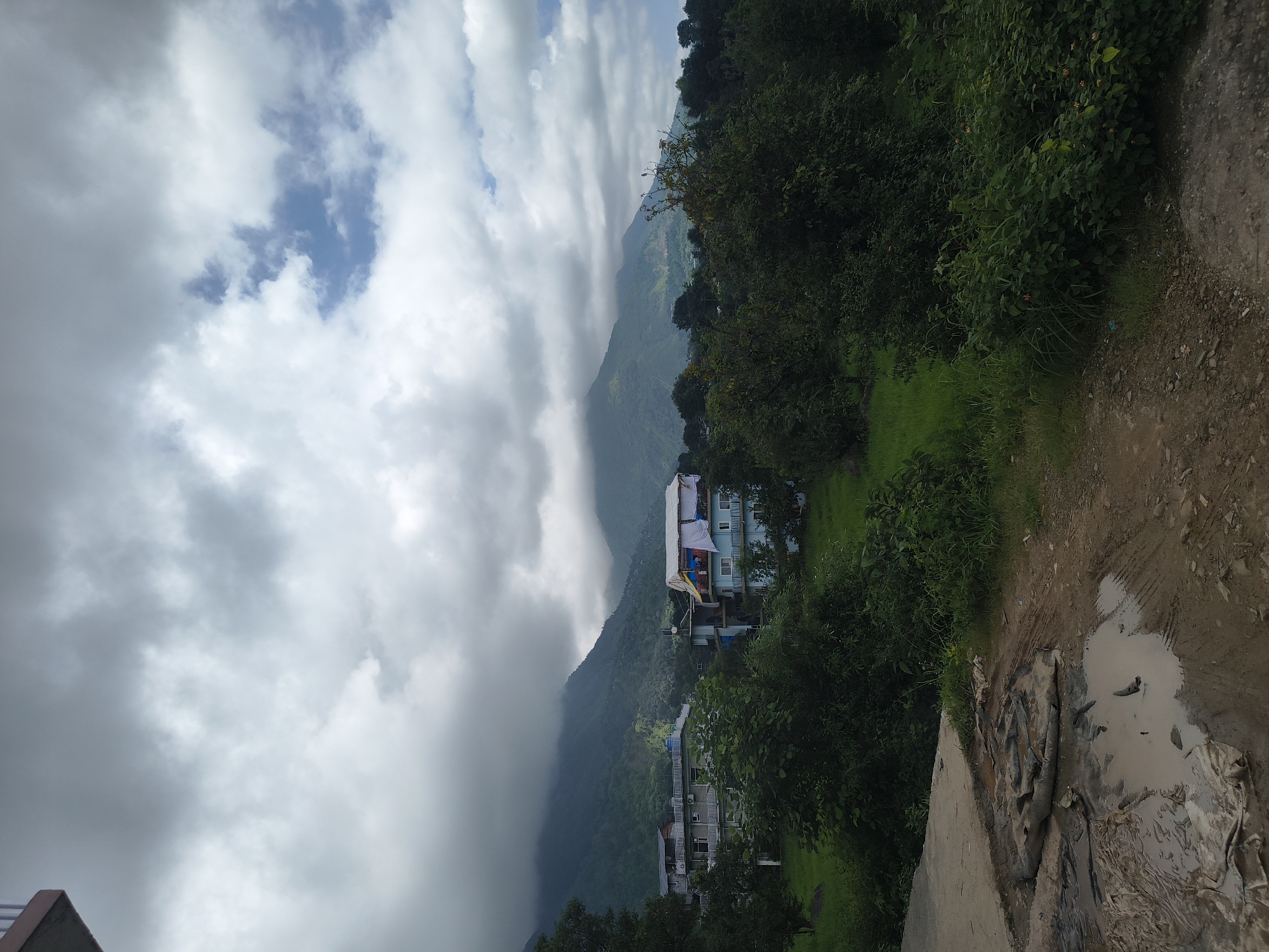 The image depicts a cloudy day in a rural or semi-rural area of Kandi, India. In the foreground, a muddy and wet area with tire tracks is visible on a dirt and concrete path. Several buildings, some appearing to be residential or guesthouses, are situated amidst lush green vegetation. The buildings have multiple stories and some have balconies. Laundry is seen drying on the roof of one of the buildings. In the background, rolling hills and mountains are covered with trees and shrouded in clouds, with patches of blue sky visible. The overall scene suggests a natural environment with human habitation.