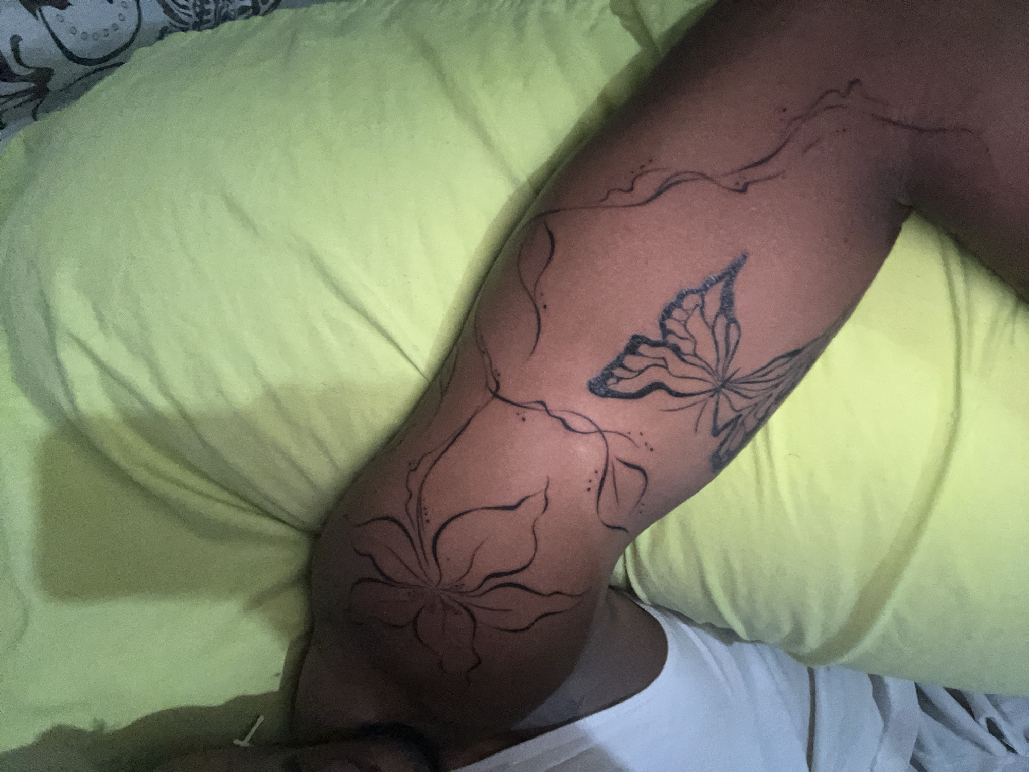 A person with a dark skin tone has their arm resting on a lime green pillow. The arm has a tattoo of a large flower with multiple petals and a butterfly with intricate wing patterns. There are also thin, swirling lines extending from the flower and butterfly, creating a vine-like design. The person is wearing a white t-shirt. The background is blurred and out of focus. No specific location in La Asuncion, Venezuela can be identified from the image.