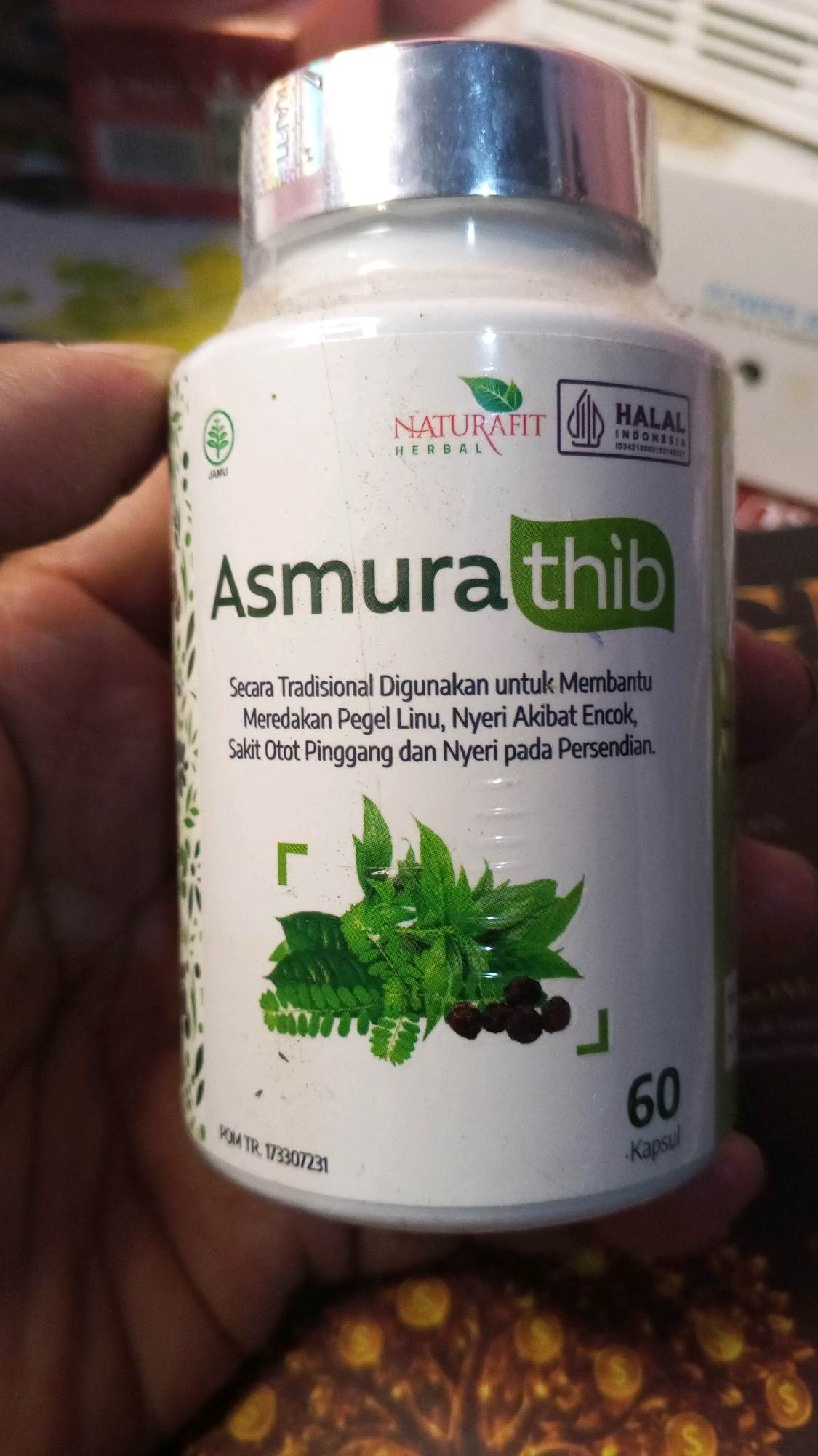 A white bottle of "Asmura thib" herbal supplements is held in a person's left hand. The bottle has a silver cap and features green lettering and leaf graphics. Text on the bottle indicates it is "Naturafit HERBAL," "HALAL INDONESIA," and lists indications such as relieving aches, pains from rheumatism, back pain, and joint pain. A green square symbol is present to the left of some plant imagery. The bottom of the bottle shows "POM TR. 173307231" and "60 Kapsul." The background is blurry, showing patterned surfaces. The context of Semarang, Indonesia, is not visually evident from the image.