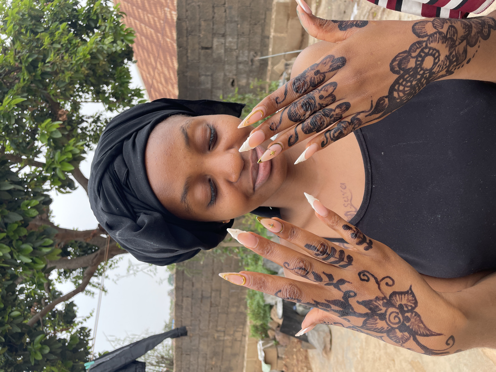 A woman with brown skin is shown from the chest up. She is wearing a black head covering and a black tank top. Her eyes are closed. She is facing forward with her hands raised, palms toward the camera. The backs of her hands are decorated with dark-colored henna designs. She has long, artificial nails with white tips and some gold accents. The background includes a brick wall and foliage, indicating an outdoor location that is in the city of Jos, Nigeria.