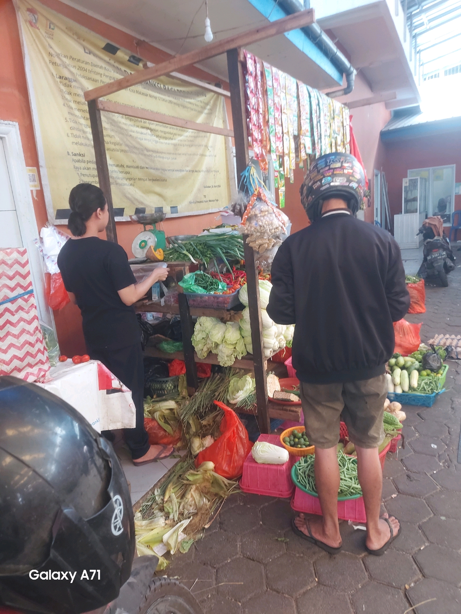 A street-level outdoor market scene is depicted in Makassar, Indonesia. Two individuals are visible. In the foreground, a person wearing a black motorcycle helmet is facing away from the camera, dressed in a black jacket and khaki shorts, with dark-colored flip-flops. To the left, a woman with dark hair pulled back is wearing a black t-shirt and black pants, facing towards a display of produce. The market stall features an array of fresh vegetables, including cabbages, green beans, chili peppers, onions, and cucumbers, displayed in baskets and crates. Behind the woman, a large yellow banner with text in Indonesian is hanging. The ground is paved with hexagonal bricks, and a motorcycle is parked in the background. The overall scene suggests a typical day at a local market.