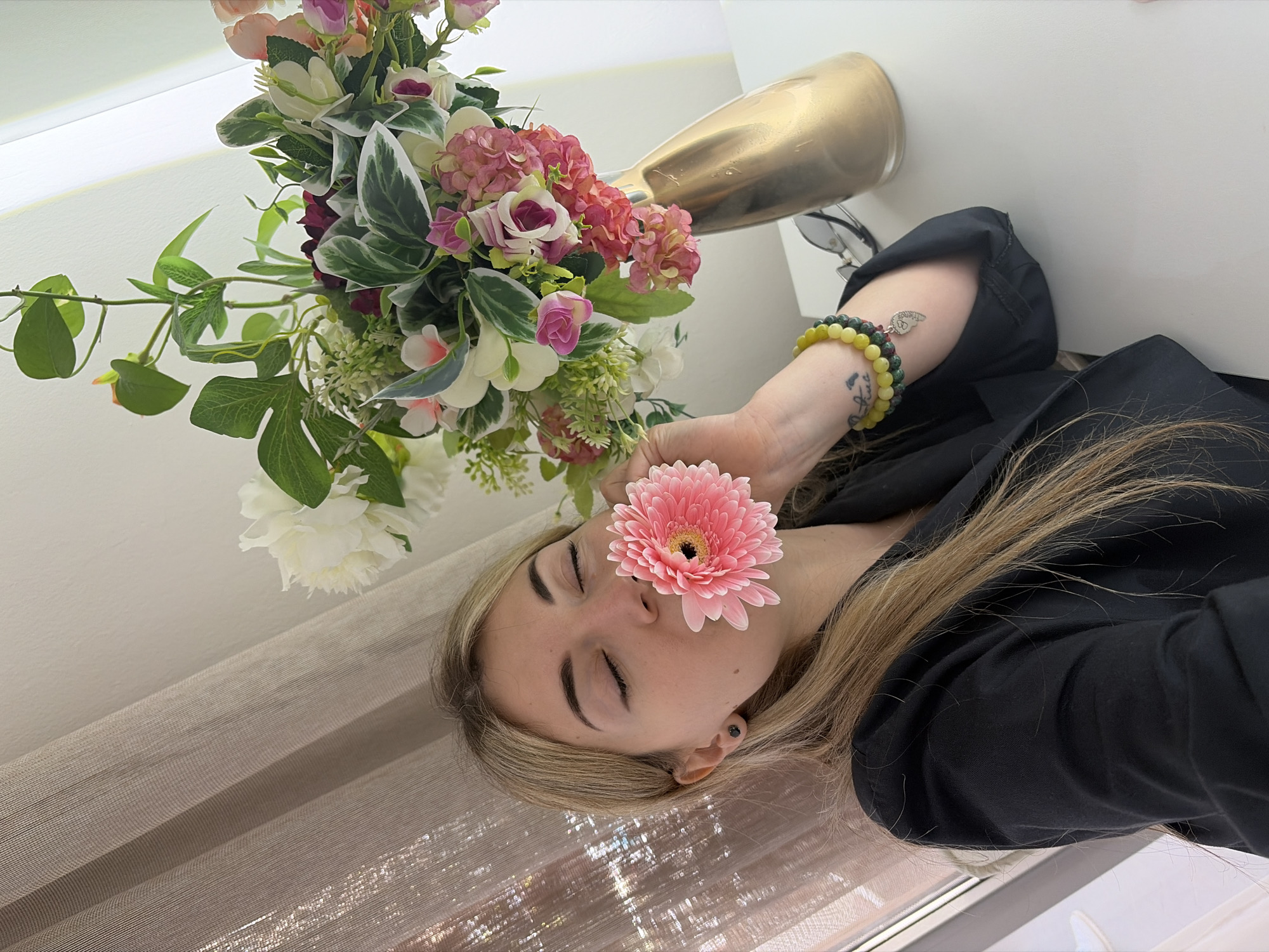A woman lies on her side with her eyes closed in an indoor setting. A pink gerbera daisy covers the middle of her face. She has blonde hair, wears a black blazer, and has a beaded bracelet on her right wrist. A bouquet of flowers in a golden vase is positioned near her head. The scene is illuminated by daylight coming through a window, and a shimmery beige curtain is visible in the foreground. There is no information to suggest the location is in Budva, Montenegro.