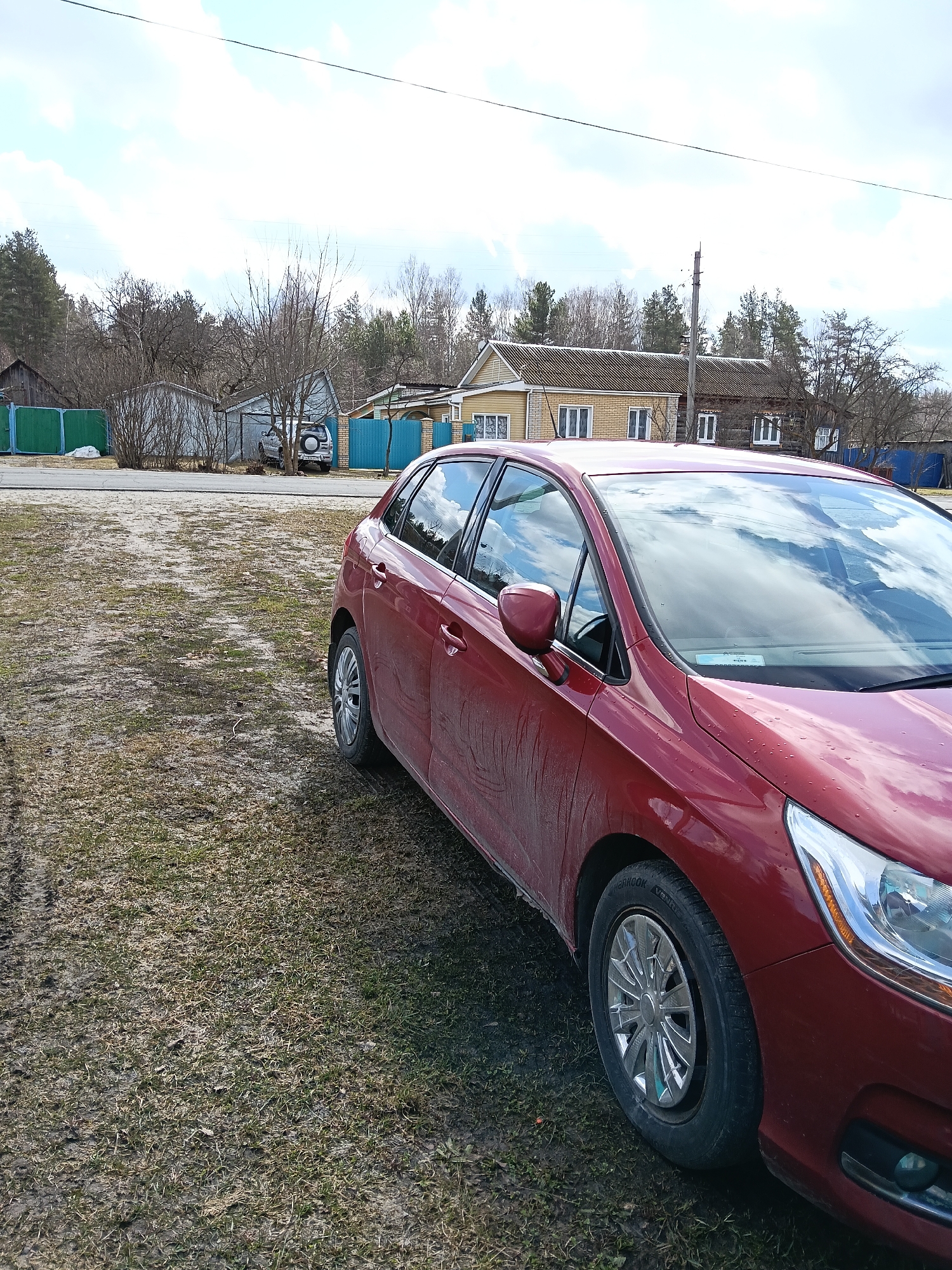 The image depicts a red Citroën C4 parked on a patch of dry grass and mud, with a dirt road and residential buildings in the background. The scene suggests a rural or semi-rural setting. The houses are characterized by wooden and brick construction with various fences, and a car is visible in the background. The cloudy sky suggests a daytime setting. The location is stated to be in Красный Маяк, Russia.