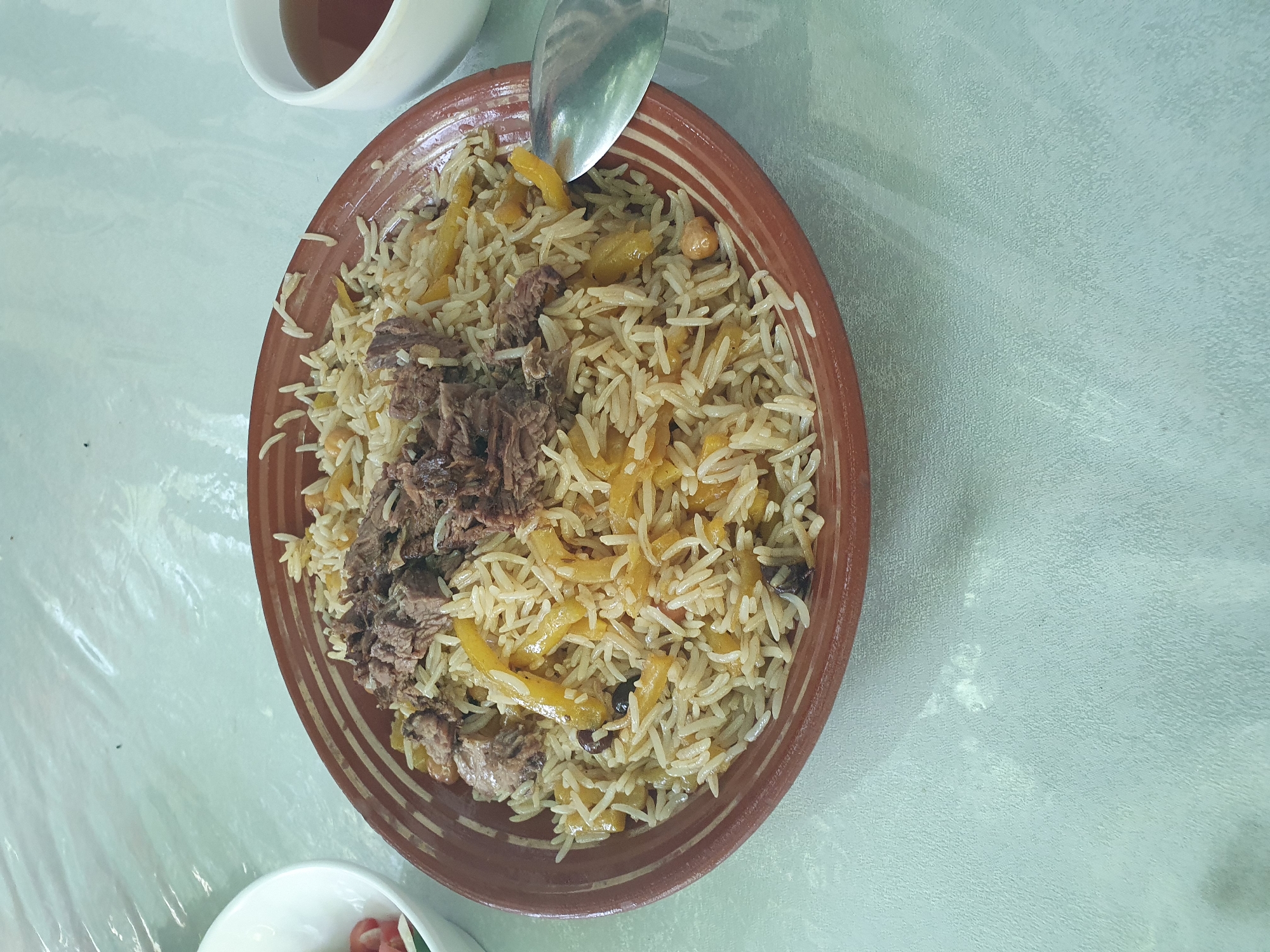 A close-up, overhead shot shows a plate of pilaf, a traditional Uzbek dish, at a restaurant in Tashkent, Uzbekistan. The pilaf, served in a wide, shallow earthenware bowl with a distinctive striped rim, is topped with shredded meat. The rice is interspersed with yellow carrot slices and chickpeas. A silver spoon rests on the right side of the plate. To the left of the pilaf, a small white bowl contains a salad with visible tomato pieces. In the upper right corner, a white teacup holds a dark liquid, likely tea. The dishes are placed on a light blue, textured tablecloth. The scene is dimly lit, and the overall impression is of a casual meal setting.