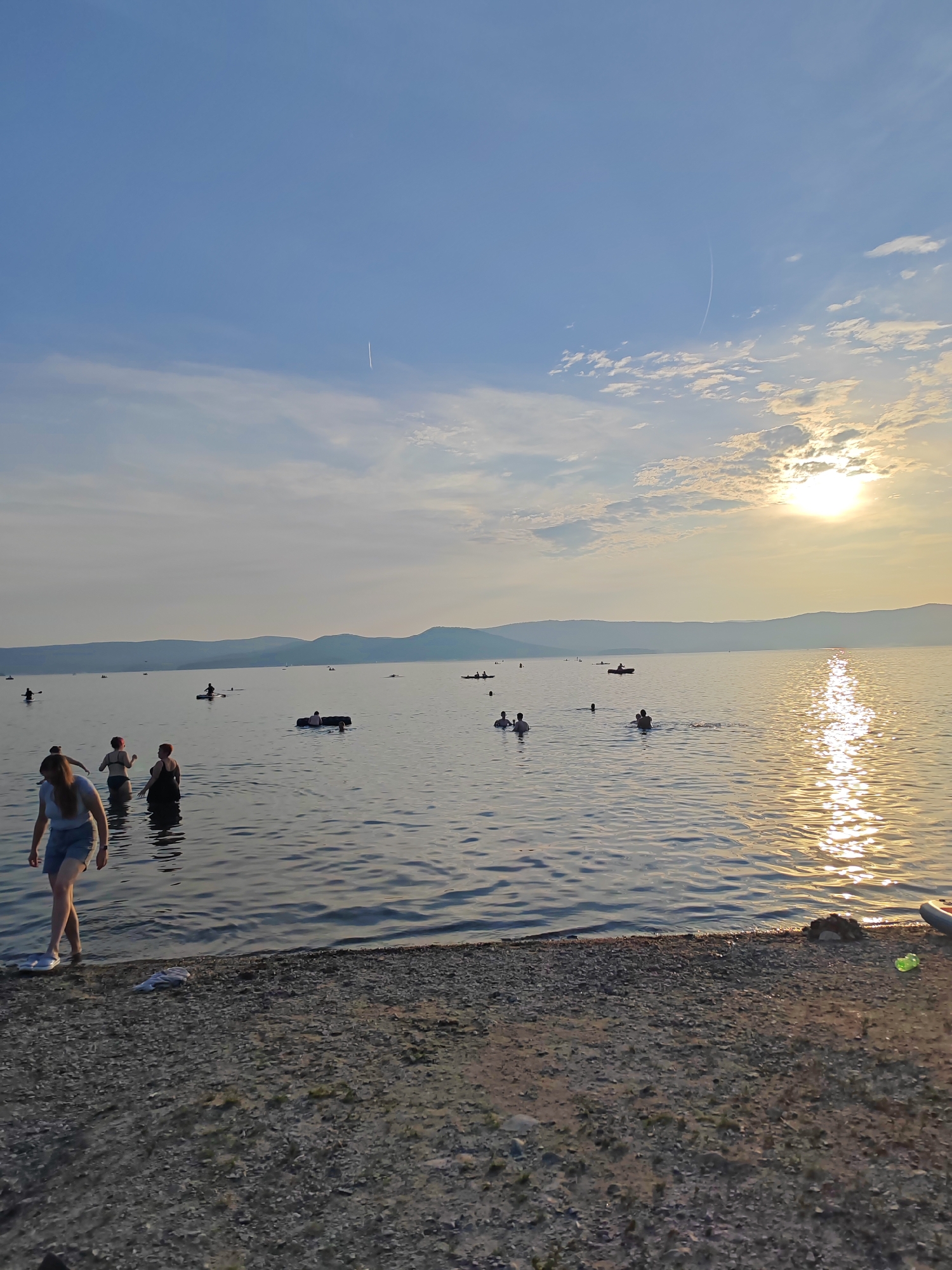The image captures a serene scene at the shore of Lake Turgoyak, Russia, during sunset. The sky is a soft blue, with wispy clouds scattered across it, and the sun's golden rays create a shimmering reflection on the calm water. In the foreground, a person is walking towards the water on a pebbly beach. Further out in the lake, several individuals are enjoying the water, some swimming, while others are on kayaks or inflatable rafts. In the distance, a range of low hills forms a gentle silhouette against the horizon, adding to the tranquil ambiance of the natural landscape.