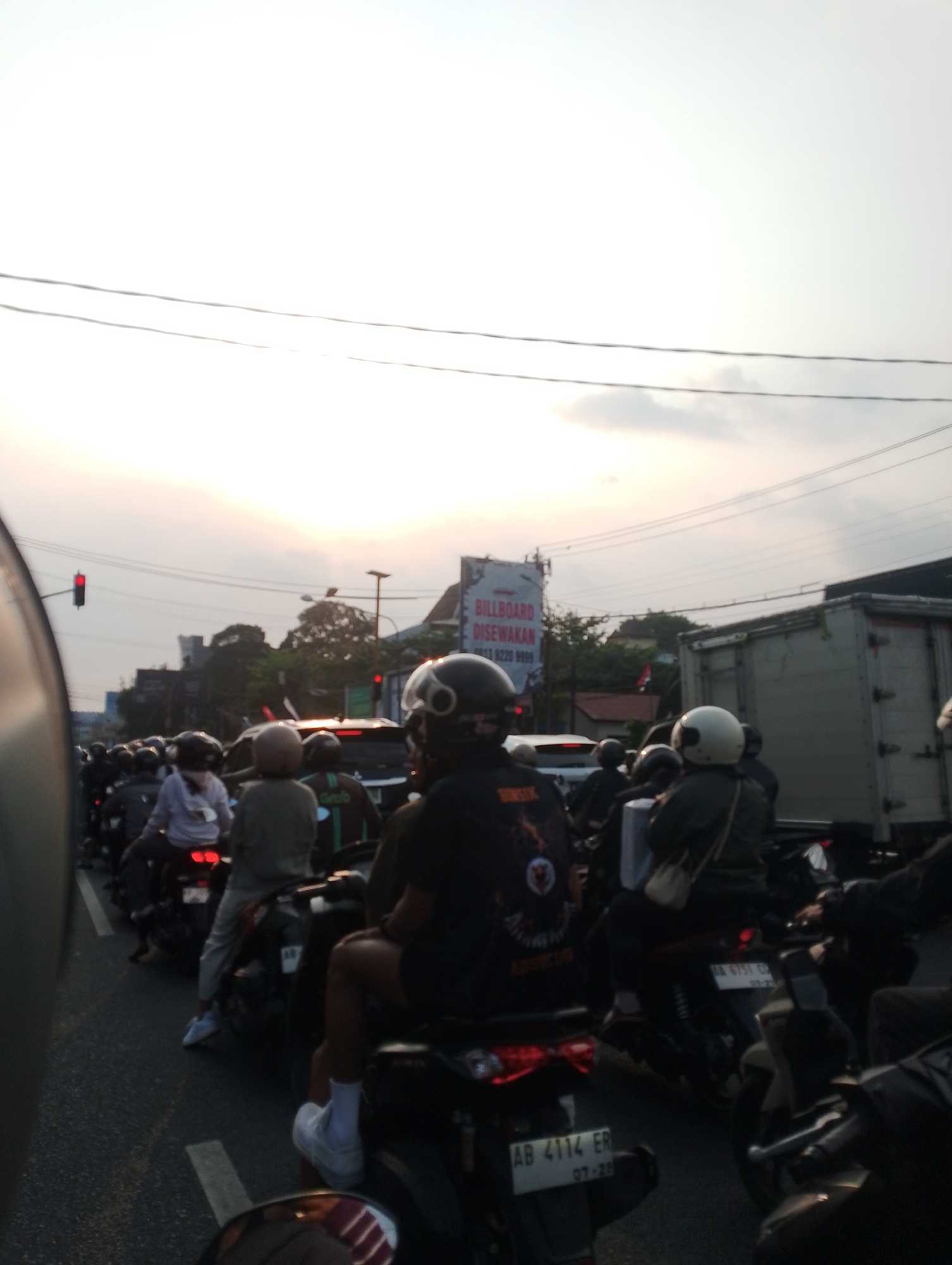 The image depicts a traffic jam on a road in Depok, Indonesia, during what appears to be sunset. Numerous motorcycles and a few cars are visible, with most people wearing helmets. The sky is overcast with some sunlight breaking through. A traffic light is red in the distance, contributing to the congestion. Billboards and buildings line the side of the road. The scene suggests a typical urban commute in Indonesia.