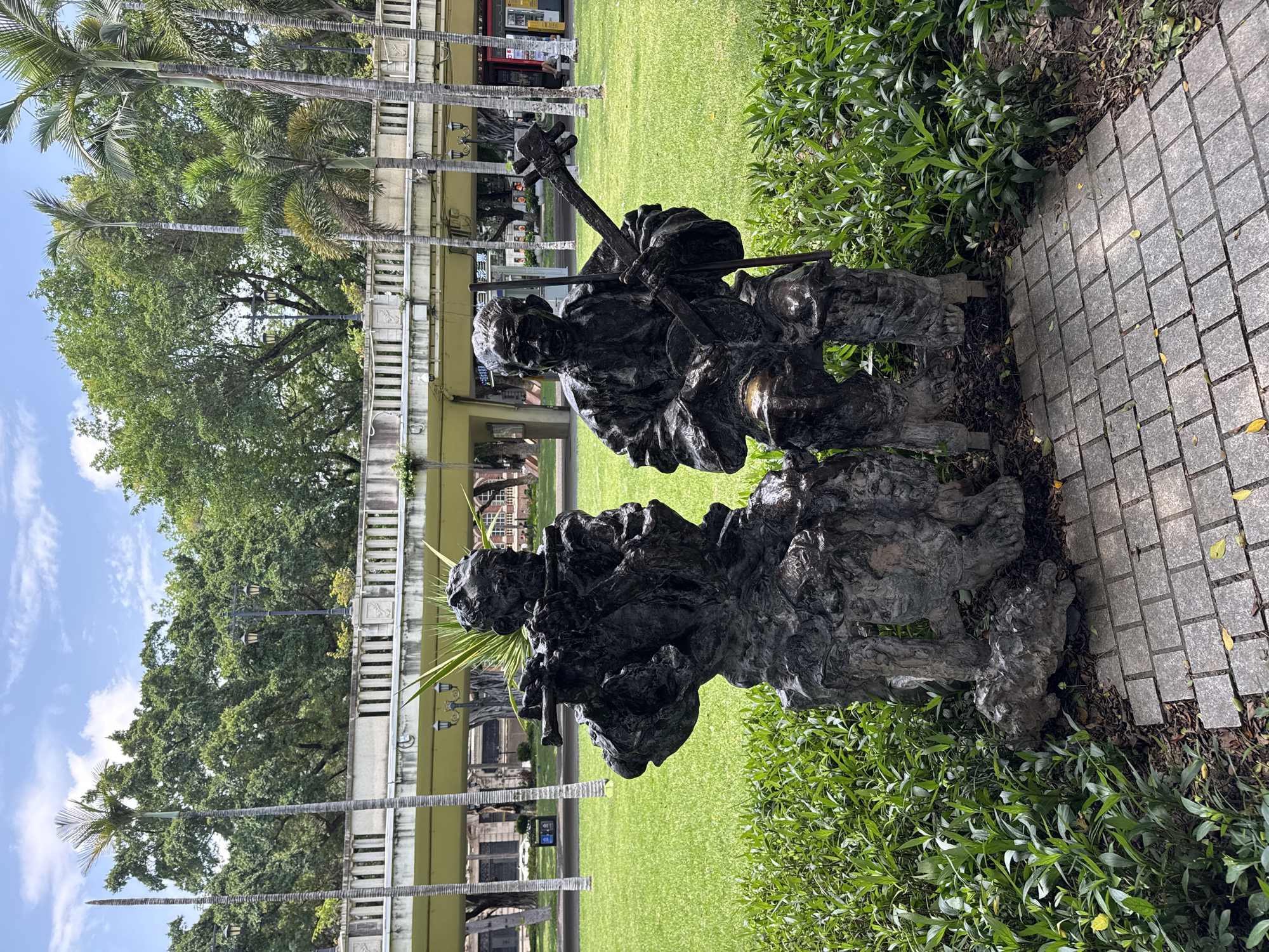 A bronze statue of two seated musicians is located in Liwan District, China. One musician holds a musical instrument with a long neck and a bow, while the other holds a flute. They sit on a stone-like base surrounded by green bushes, with a gray-tiled pathway to the right. The background features a green lawn, palm trees, and a light-yellow building with a bridge-like structure and pillars.