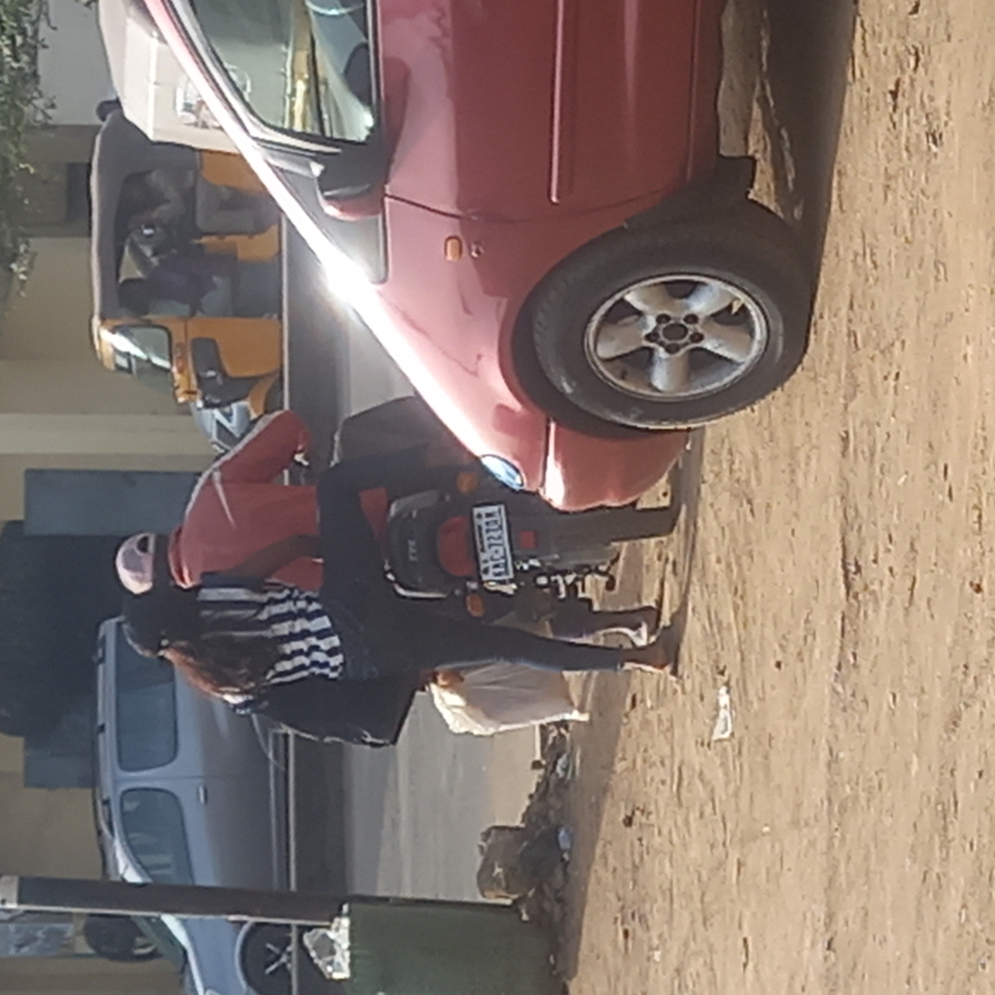 The image captures a street scene in Dadi Riba, Nigeria, under bright daylight. A red hatchback car is parked prominently on the right, with its front end and a portion of its right side visible, reflecting strong sunlight. Its silver multi-spoke wheel is clearly seen.
In the mid-ground, a person, likely female with long dark hair, is seated on a black TVS motorcycle. She wears a pink helmet, a red long-sleeved top over a black and white striped top, and blue leggings. She appears to be barefoot or wearing minimal footwear. The motorcycle's license plate, "KBL-2720-KM," is clearly visible at the rear.
Behind the red car, a yellow auto-rickshaw (often called a Keke Napep in Nigeria) is partially visible, with a person seated inside. Further to the left and behind the motorcycle, part of a silver or light-colored car is also parked.
The ground is unpaved and dusty, with visible dirt, gravel, and some scattered litter or debris, particularly in the lower left foreground near a dark green refuse bin. The background consists of indistinct buildings or structures and some greenery. The overall impression is a bustling urban or semi-urban environment.