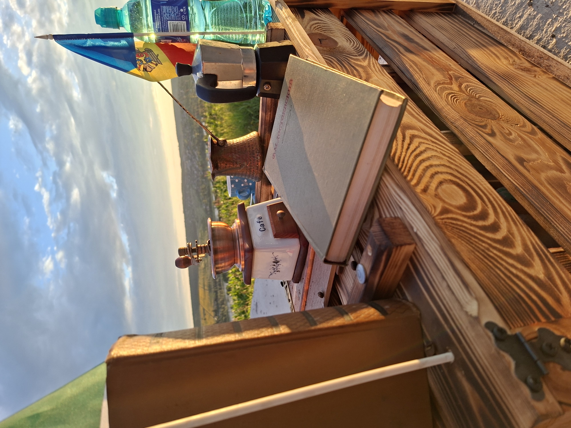 The photograph captures a close-up view of a wooden crate table outdoors, with a scenic overlook of the city of Sociteni, Moldova, in the background. The setting appears to be during golden hour, as indicated by the warm, soft lighting illuminating the scene.
On the table, several objects are arranged: a large, open book with a brown cover is propped up on the left. To its right sits a white ceramic coffee grinder with a copper top, displaying the word "Cafe" in black lettering. Beside the grinder, a small copper pot, likely a cezve for making Turkish coffee, is visible. A silver Moka pot, a polka-dot mug, and a bottle of clear liquid with a blue cap are also present. A small Moldovan flag, with its blue, yellow, and red colors and coat of arms, is planted in the arrangement. In the foreground, a closed book with a light brown cover lies on the crate.
The background reveals a gently sloping landscape with green fields and scattered buildings, characteristic of a town or village. The sky is partly cloudy, with a blend of blue sky and white and grey clouds, suggesting a pleasant evening. The overall scene evokes a sense of leisure and relaxation, possibly a picnic or a moment of quiet contemplation in a picturesque location.