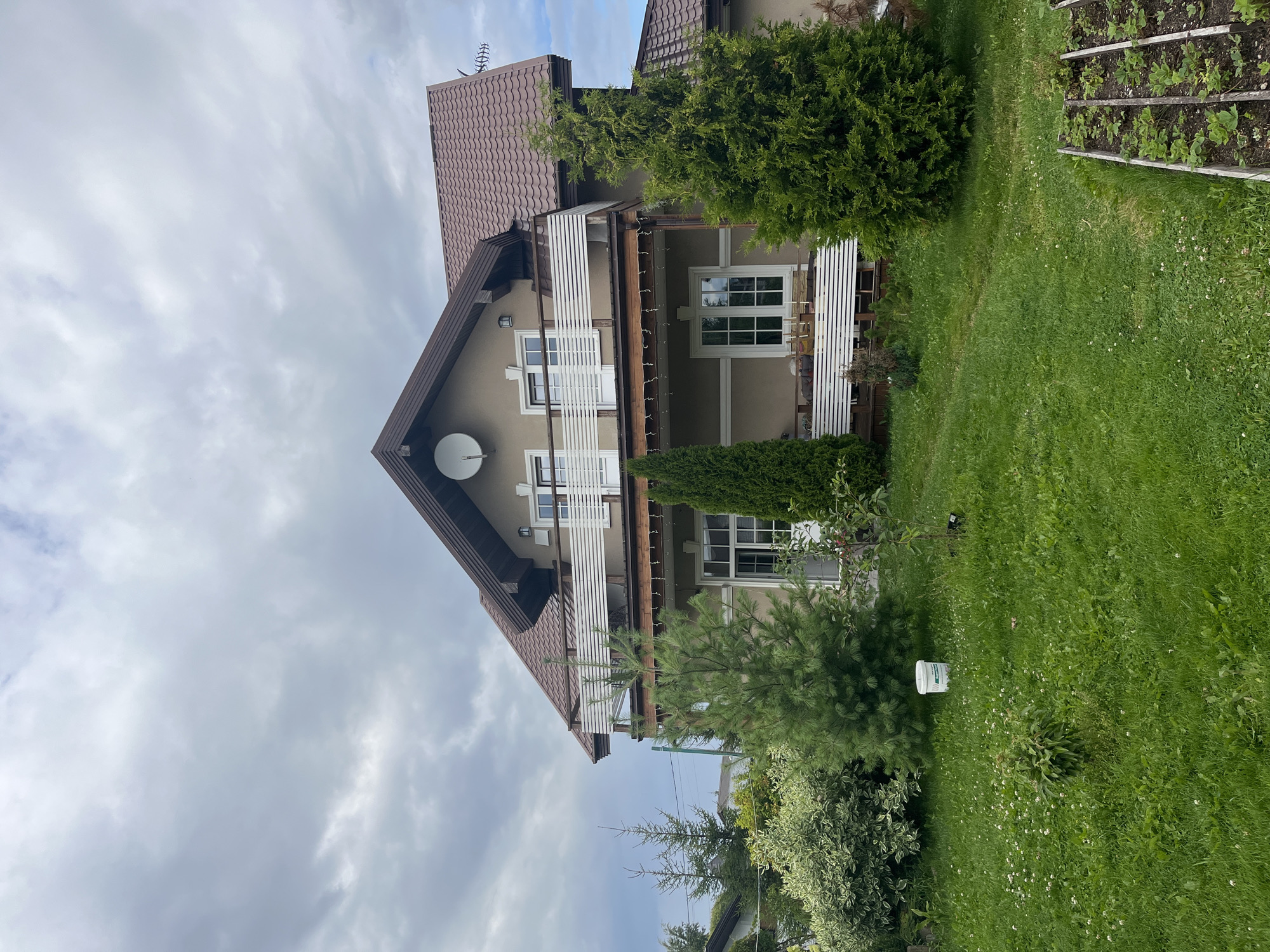 A wide, eye-level, outdoor, medium shot captures a two-story house with a brown, tiled roof, beige siding, and white-trimmed windows and doors, on a cloudy day in Vladimirovka, Russia. A white, slatted balcony extends across the second story, and a similar slatted porch is visible on the first. Two green evergreen trees flank the front of the house, with a smaller pine tree in front of the left evergreen and a thin, tall evergreen tree to the left of the porch. A white bucket sits in the grass in front of the pine tree. The foreground features a lush, green lawn with scattered white flowers, and a wooden raised garden bed with small green plants is visible in the bottom right corner. A satellite dish is mounted on the roof near the peak. The sky is heavily clouded with muted gray tones.