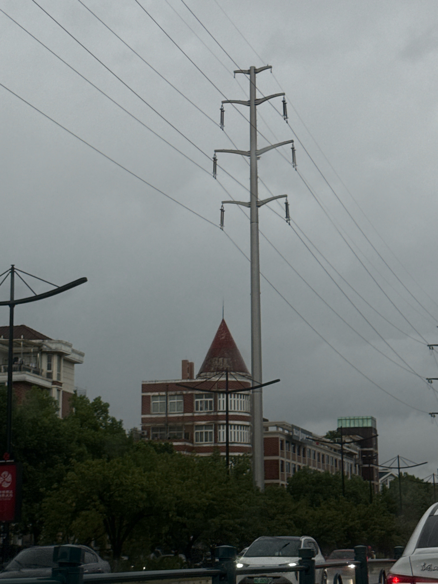 The image depicts a cloudy day in Yiwu, China. A large electricity pylon dominates the center-right of the frame, with several high-voltage power lines stretching across the sky. In the background, several buildings are visible, including a brick building with white-trimmed windows and a distinctive conical red roof. Trees with green foliage line the foreground, partially obscuring a street with several cars. The overall scene suggests an urban environment with infrastructure and residential or commercial buildings.