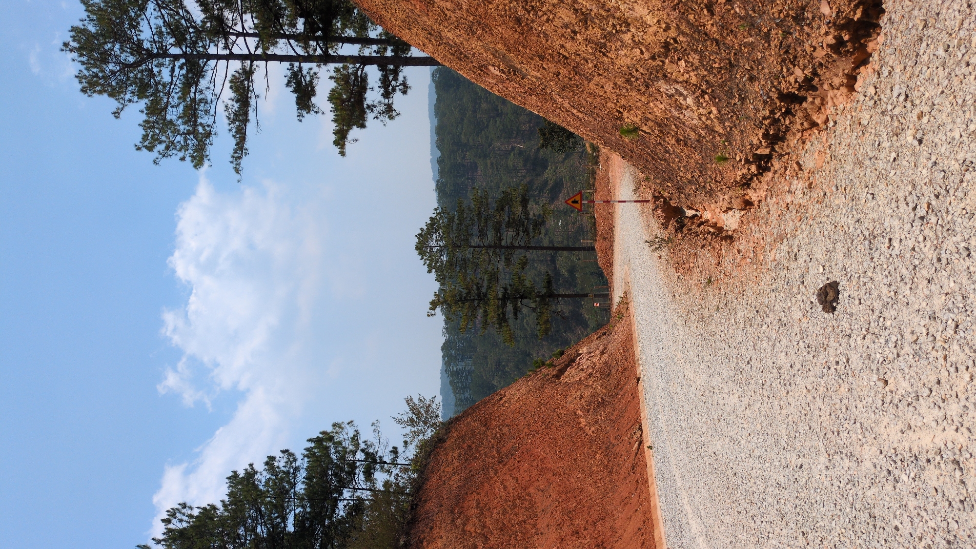 A gravel road cuts through a landscape in Lạc Dương, Vietnam, flanked by red-brown soil banks. The road, angled diagonally in the frame, leads to a yellow warning sign further along. Pine trees are visible on the banks, extending towards a blue sky with scattered clouds. In the background, more forested hills rise into the hazy distance. A dark object sits on the road surface.