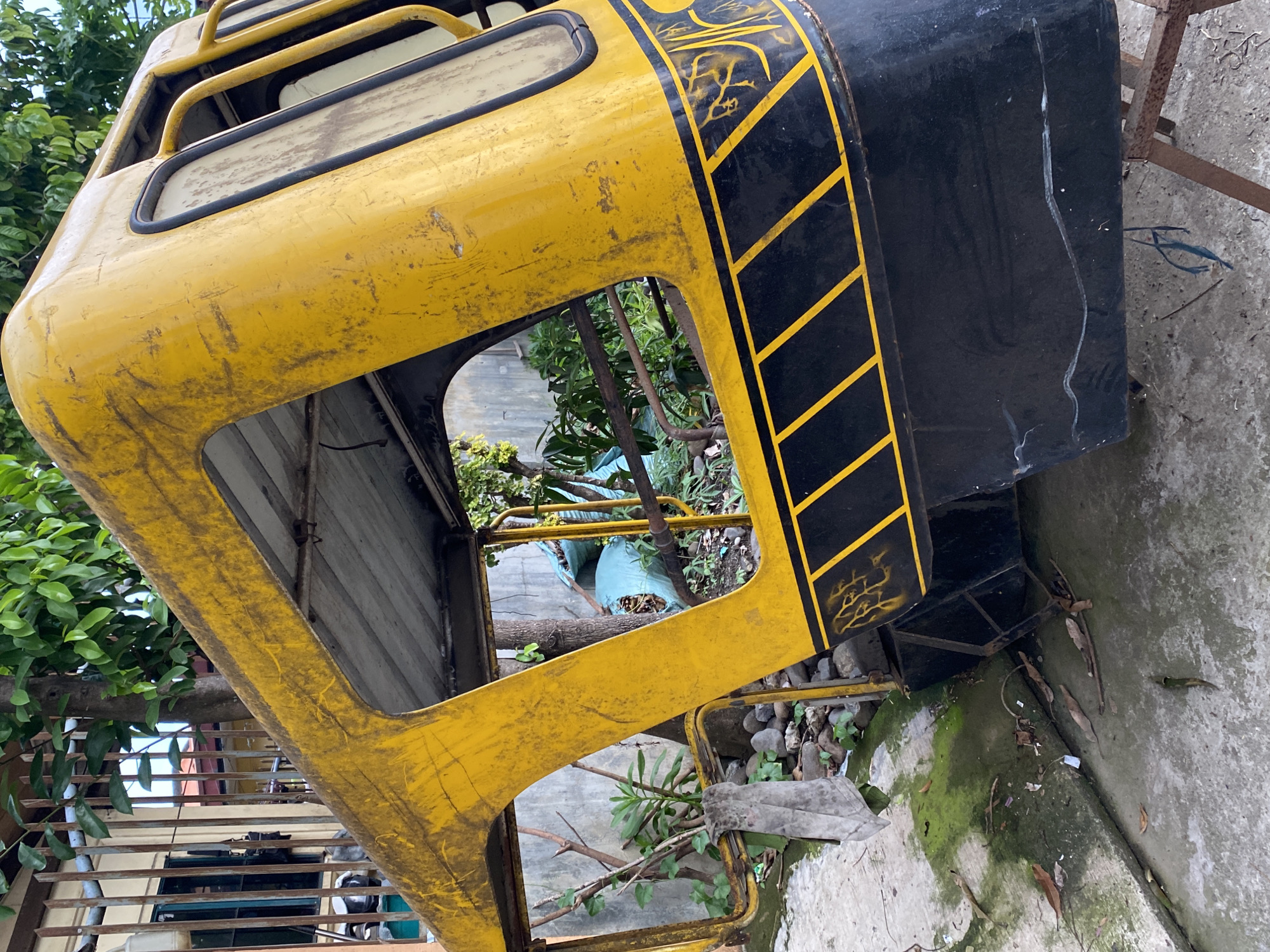 This image depicts a highly weathered, disassembled yellow and black vehicle cabin, likely from a motorized tricycle, positioned outdoors in what appears to be a residential or semi-industrial yard in Davao City, Philippines. The main subject, an inverted or tilted metal shell, is predominantly bright yellow on its upper section, heavily marked with scratches, grime, and streaks of dirt, indicating significant exposure and wear. Its lower section is matte black, also showing scuffs and minor damage, adorned with yellow diagonal stripes and some faded, abstract yellow graffiti-like markings. The structure exhibits multiple open window cutouts and exposed metal framing, with visible rust on some edges and support bars. A dirty, light-colored cloth rag hangs from a metal bar on the left side.
The vehicle shell rests on a rusty, brown metal support frame, particularly visible beneath its black base on the right. The ground beneath is concrete, stained with patches of green moss and algae, and scattered with dry leaves, small twigs, and miscellaneous debris.
In the background to the left, a light-colored building, possibly a house, is visible behind a rusty metal fence or grille, partially obscured by green foliage and a tree trunk. A dark-framed window with a teal tint is visible on the building. To the right, behind the yellow shell, a gray concrete wall is present, with various green plants and bare tree branches growing in front of it. A large, light-blue tarp or bag, possibly containing refuse or garden waste, is noticeable near the wall. No people or animals are present in the scene, and no actions or interactions are depicted; the image captures a static outdoor setting with discarded or stored items.