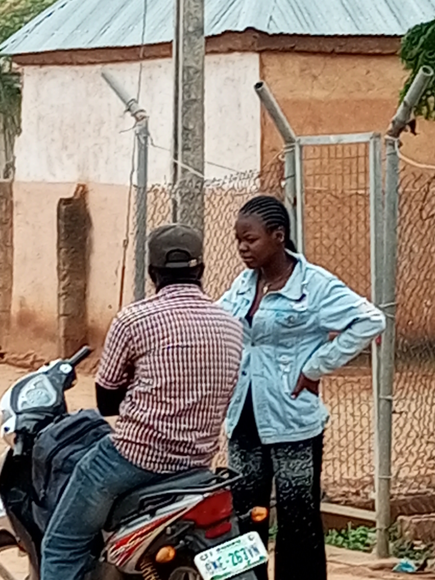 The image shows two individuals, a male and a female, standing outside a building in Gombe, Nigeria. The male, seated on a motorcycle, is wearing a red and white striped shirt and blue jeans, with a dark baseball cap on his head. The female, standing next to him, is wearing a light blue denim jacket over a dark top and black patterned trousers. She has her left hand on her hip. The background features a building with a corrugated metal roof and a textured wall, partially obscured by a chain-link fence. A motorcycle is visible in the foreground, with a license plate that reads "KAE-263VN". The overall scene appears to be a typical street view in Gombe.