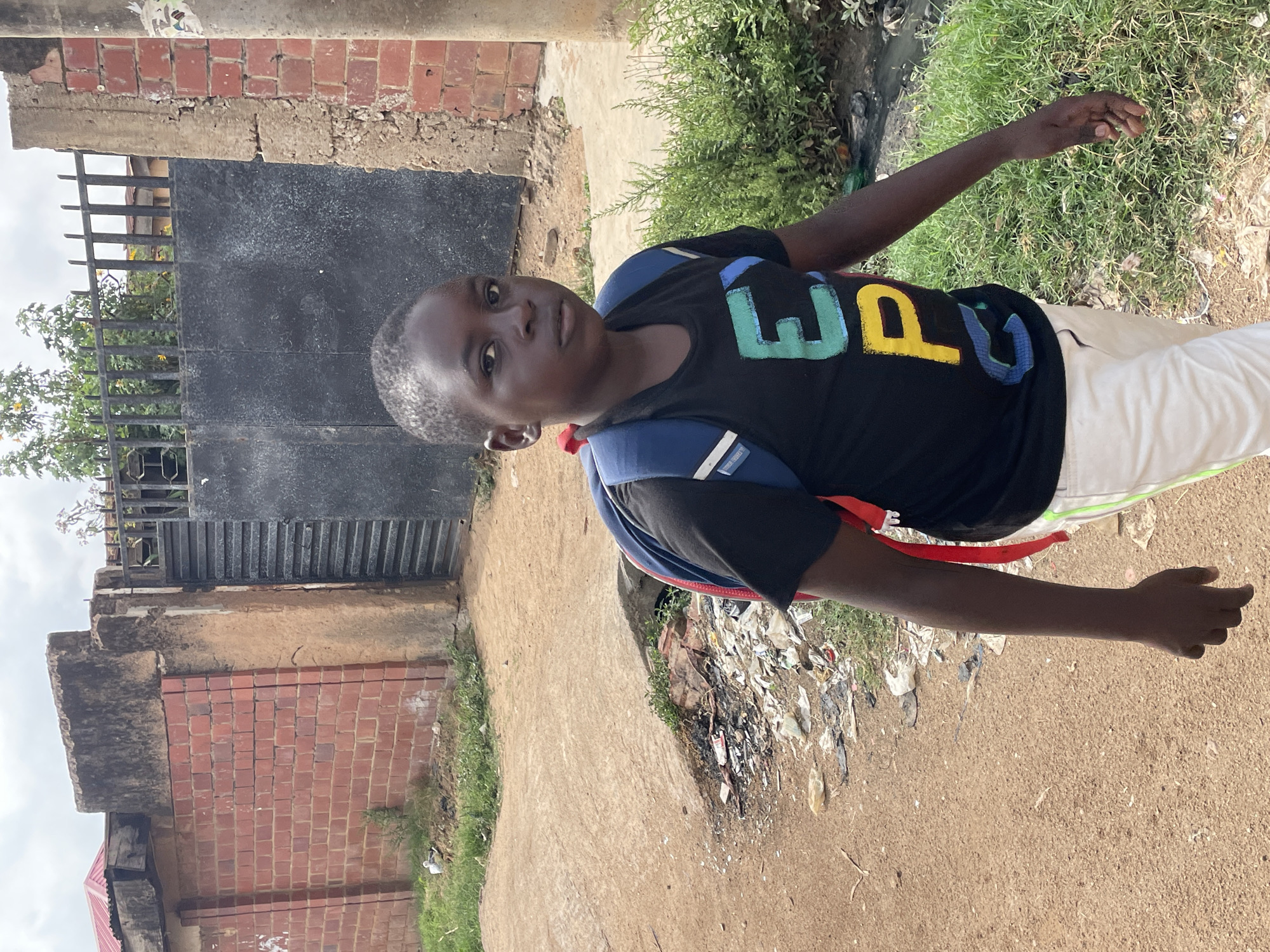 A young boy with short hair stands outside next to a dirt road with litter and vegetation. He wears a black shirt with a colorful word design and a blue and red backpack. A brick wall and a metal gate are visible behind him, with a cloudy sky above. The location is Jos, Nigeria.