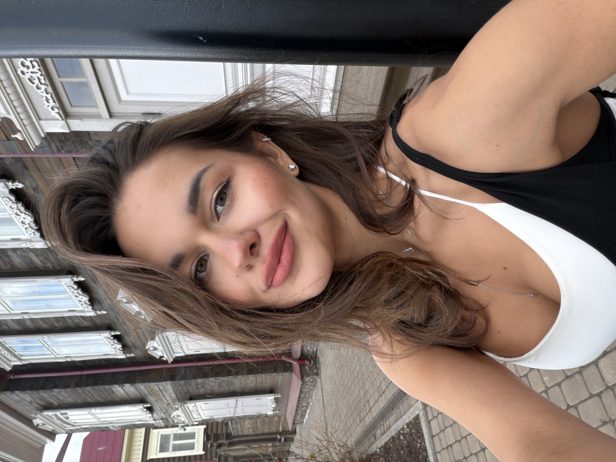 A selfie photograph shows a woman with shoulder-length brown hair, fair skin, and light brown eyes. She is smiling slightly, wearing a black and white tank top, a silver necklace, and earrings. The background shows a street in the city of Krasnoyarsk, Russia. A wooden building is visible on the left side of the image. The sidewalk is made of light gray pavers. There is also a black bar in the background. The photograph is taken at an angle.