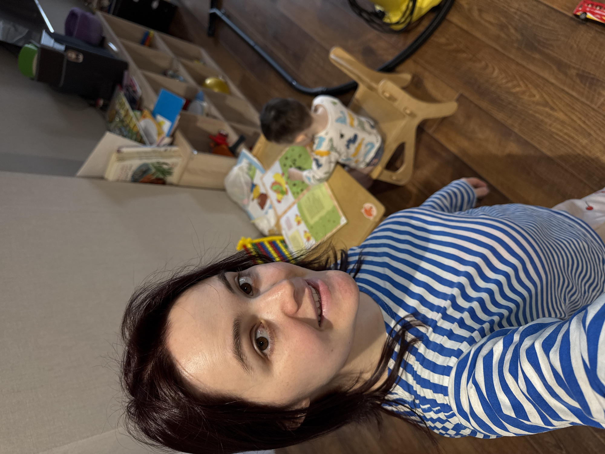 A woman with dark hair and a blue and white striped shirt is lying on a wooden floor. A young child in a patterned onesie is kneeling on the floor looking at a book. The book is on a small wooden table next to a wooden rocking horse. In the background is a wooden shelving unit containing various items. The location is Izhevsk, Russia.