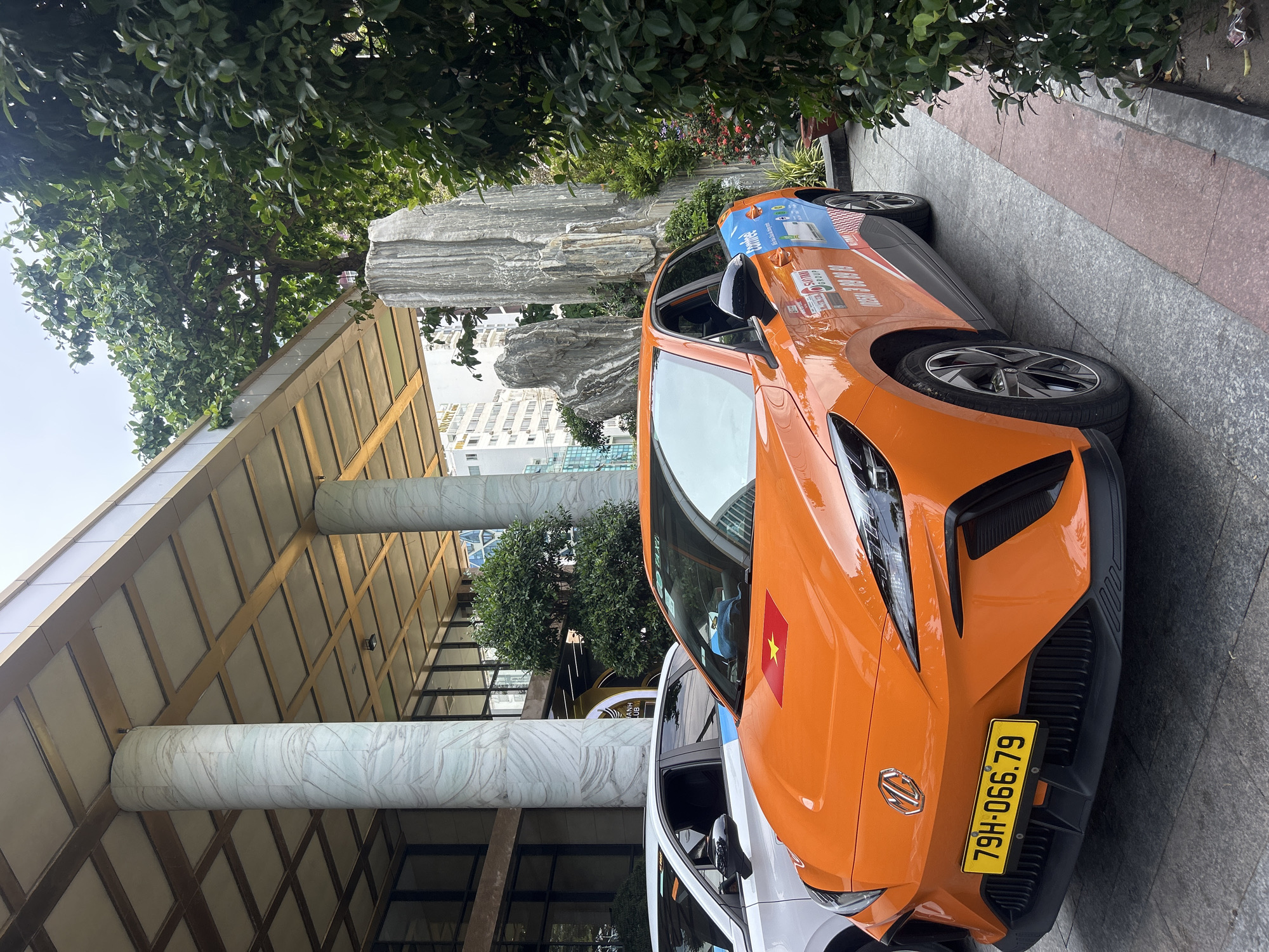 An orange MG car is parked front-first under the canopy of a building entrance during daytime in Nha Trang, Vietnam. The car, with a vibrant orange paint job, features a Vietnamese flag sticker (red with a yellow star) on its hood. Its front license plate, "79H-066.79", is yellow with black characters, typical of Vietnam. On the side of the car, partially visible decals indicate it belongs to "VINASUN GROUP" and is a "VINASUN Taxi," alongside a phone number "1055" and other Vietnamese text.
Behind the orange car, a portion of a white car is visible. The cars are positioned on a paved area, likely a driveway or drop-off zone. The building overhead has a distinctive architectural style, featuring large, round, marble-patterned pillars supporting a ceiling with a grid-like pattern and golden accents.
To the right of the cars, lush green foliage forms a dense canopy, with parts of large, natural-looking rock formations visible behind the trees and plants. In the background, modern multi-story buildings can be seen under a bright sky. The overall scene suggests an entrance to a hotel or a significant commercial establishment, blending urban architecture with natural landscaping.