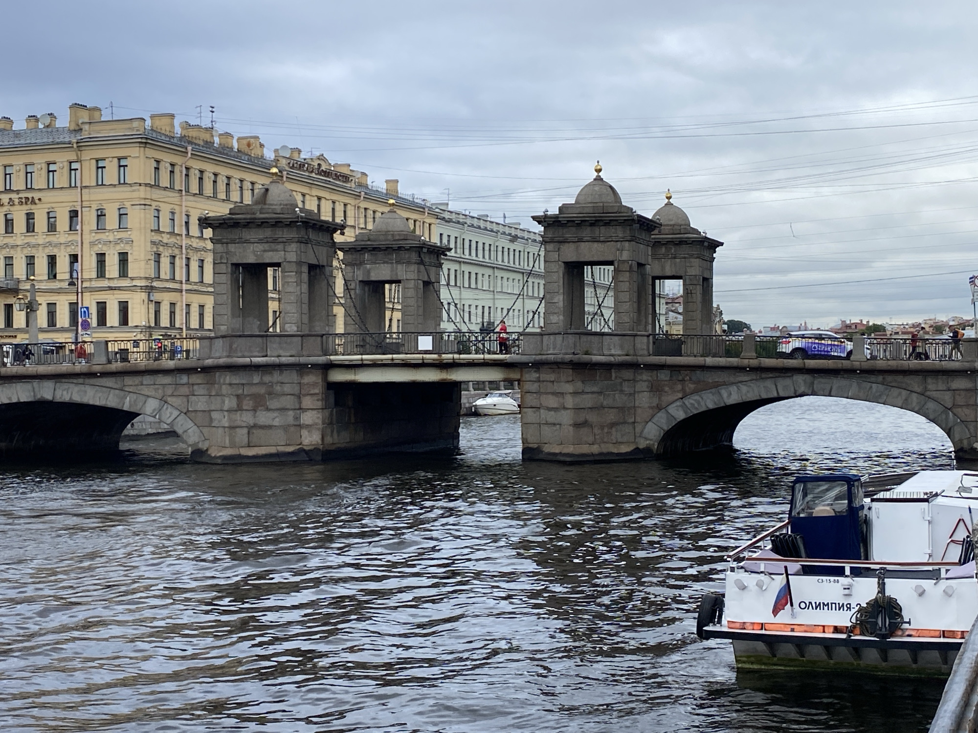 The image depicts the Bank Bridge (Lienny Bridge) in Saint Petersburg, Russia, on an overcast day. The bridge, a pedestrian suspension bridge, spans a canal and is characterized by its iconic griffin sculptures, although they are not visible from this angle. The bridge's stone piers and the arches of its lower structure are prominent. On the bridge, several people are visible walking. In the foreground, a white boat with a blue cabin is traveling on the canal. The boat has Russian flag markings and text that appears to be in Cyrillic, possibly the name of the boat. The background features classical Saint Petersburg architecture, with tall, light-colored buildings lining the canal. The sky is cloudy and grey, contributing to the muted tones of the scene. The water in the canal is rippling, indicating movement from the boat and possibly the wind.