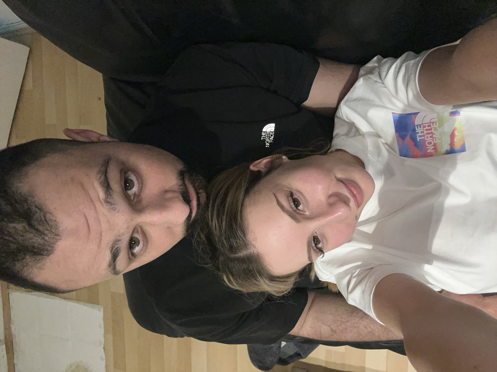 A close-up selfie taken indoors features a man and a woman lying down. The man, positioned on the left, has dark hair, a beard, and is wearing a black t-shirt with "THE NORTH FACE" logo. The woman, on the right, has blonde hair and is wearing a white t-shirt, also with "THE NORTH FACE" logo. They are lying on what appears to be a dark-colored couch. A portion of the wooden floor is visible in the background.