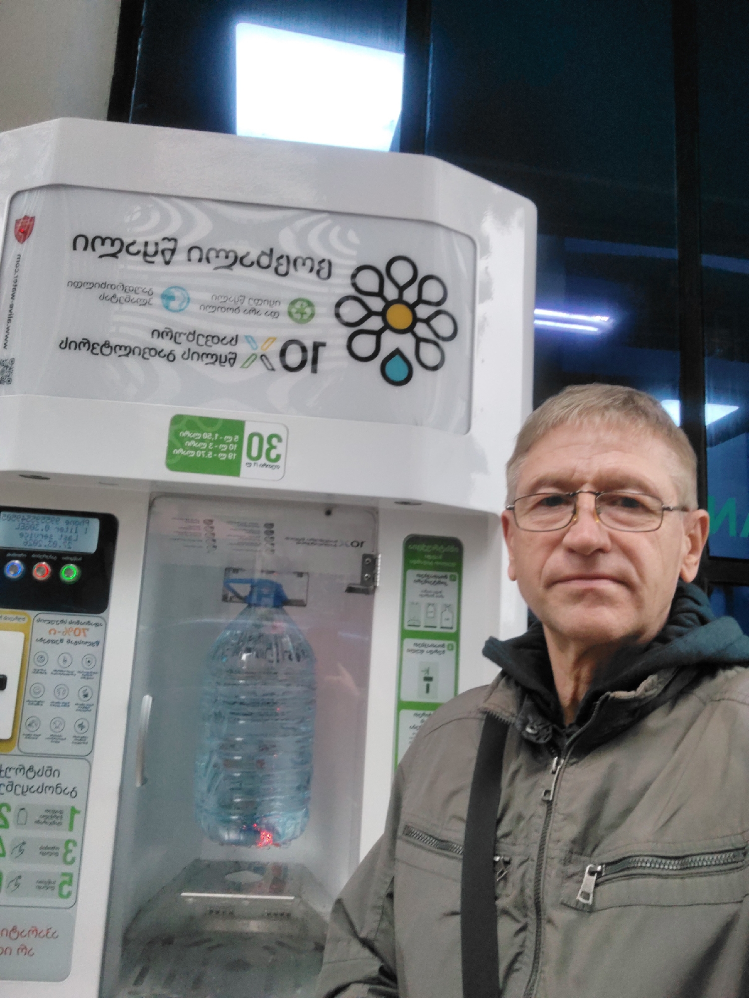 An older man with short gray hair and glasses stands facing the camera in front of a water refill kiosk with Georgian text. The kiosk has a water bottle inside a clear compartment and displays pricing information. The man is wearing a gray jacket over a dark hooded sweatshirt with a dark strap over his shoulder. The background shows windows with some interior lighting. The OCR data mentions the location, likely Batumi, Georgia, but this cannot be definitively confirmed from the image alone.