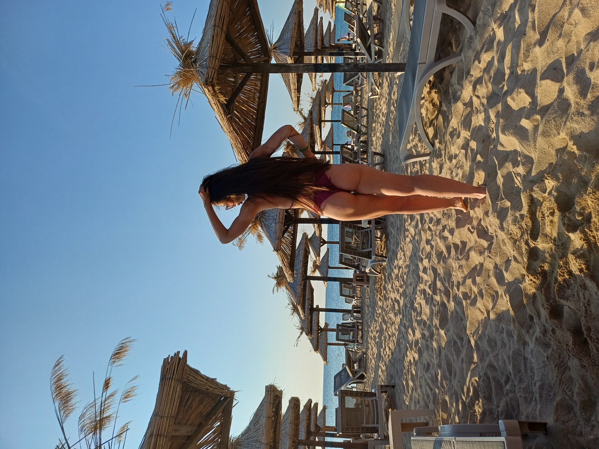 The image depicts a beach scene in Varna, Bulgaria. In the foreground, a woman with long dark hair is walking away from the camera, wearing a burgundy bikini. She has her right hand raised to her hair and her left hand on her hip. The beach is sandy, with rows of empty lounge chairs and thatched umbrellas stretching towards the sea. The sea and sky are visible in the background under a clear blue sky. The sun is casting long shadows across the sand, indicating it is likely late afternoon or early morning.