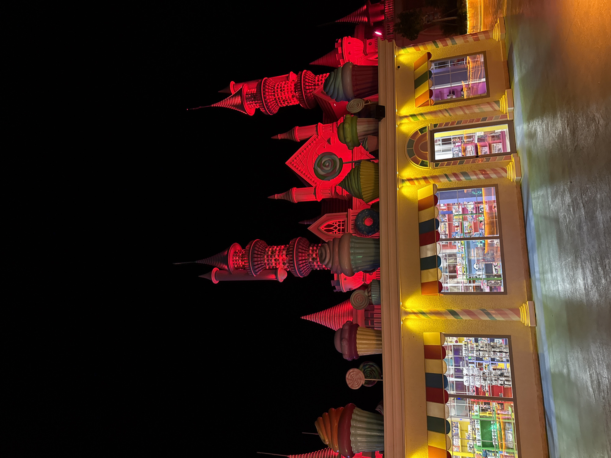 This image captures an exterior view of a candy-themed attraction at night in Bodrum, Turkey. The main structure is a whimsical building with a castle-like facade, illuminated in vibrant red lights. The foreground features a storefront with large display windows showcasing shelves stocked with candy and toys. Colorful awnings, striped columns, and oversized decorative cupcakes and lollipops adorn the storefront and roofline, contributing to the playful, fantasy-like atmosphere. The ground in front of the store is a smooth, reflective surface. The background is dominated by the dark night sky and the illuminated red castle.