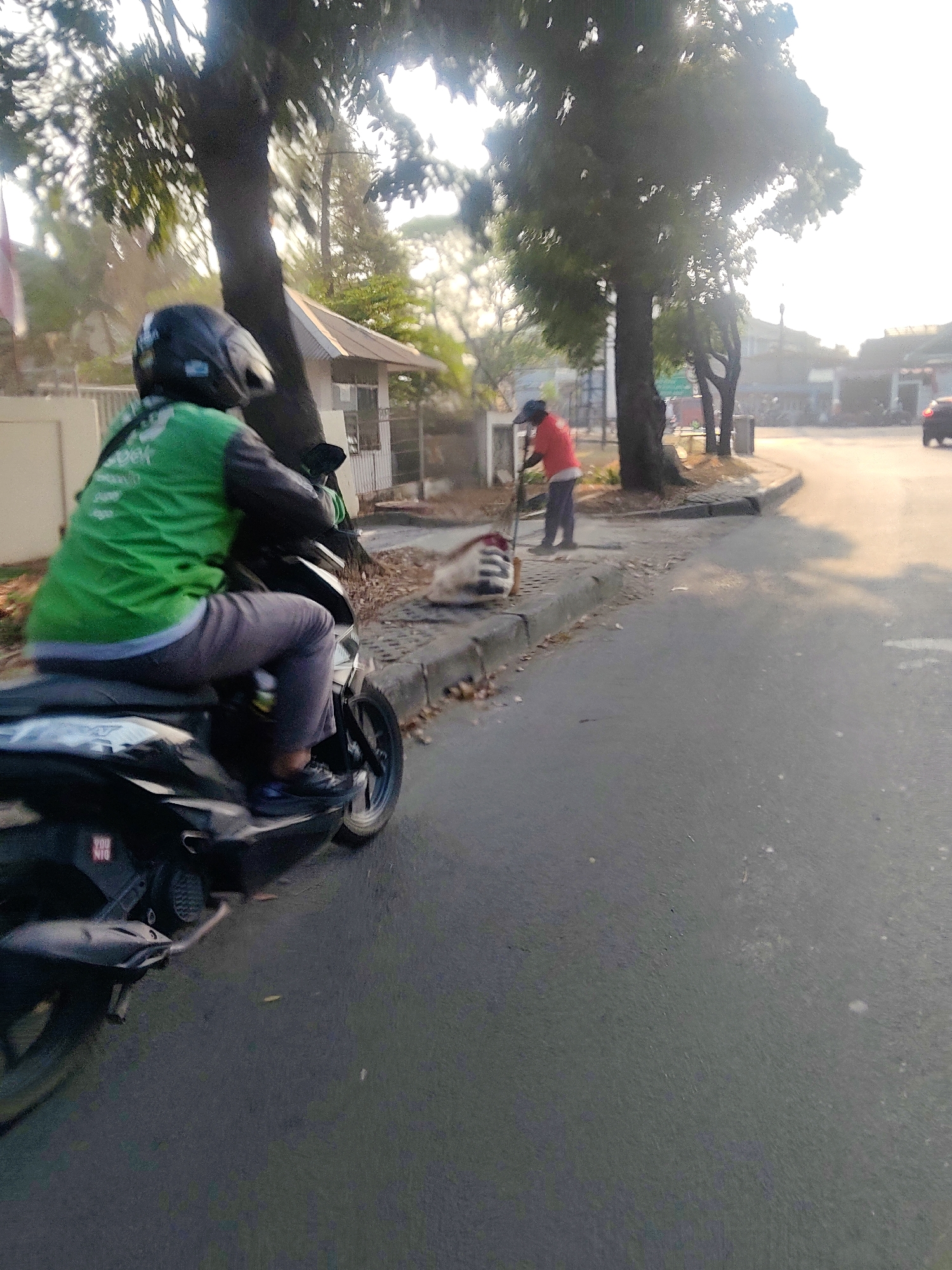 A person wearing a green jacket and a black helmet is riding a black motorcycle on a paved road. They are moving towards the viewer's left. In the background, another person wearing a red shirt and dark pants is sweeping the sidewalk with a broom. A pile of leaves is near the person sweeping. The scene appears to be an urban environment with trees and buildings visible. The lighting suggests it is daytime, possibly early morning or late afternoon due to the low angle of the sun. The location is specified as Mekarmukti, Indonesia.