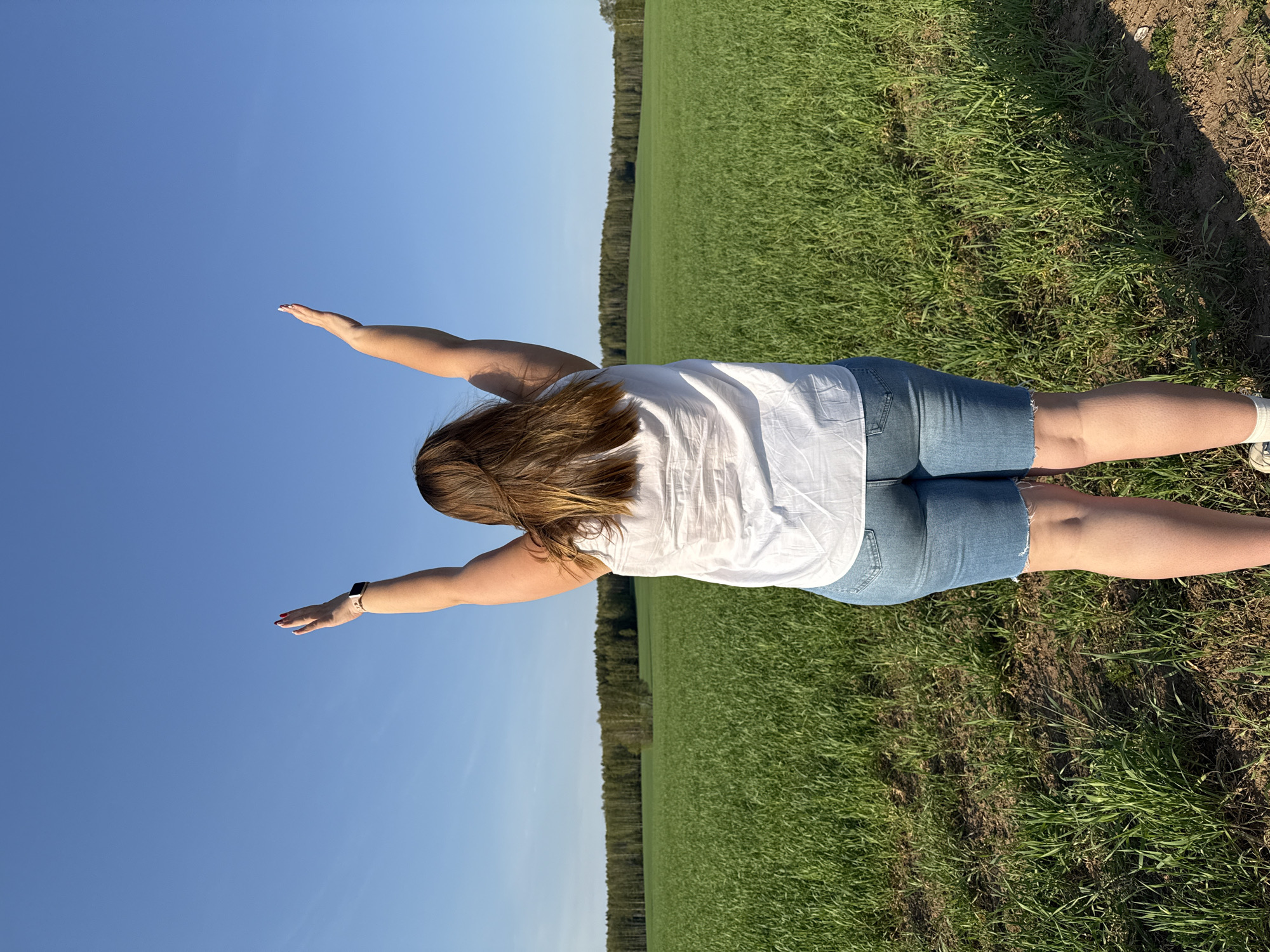A back-facing, full shot of a woman in a white t-shirt and denim shorts standing in a field near Шумиха, Russia. Her arms are outstretched. The sky is clear blue. A line of trees is visible in the background where the field meets the horizon. The field is predominantly green with brown patches of dirt. She wears a watch on her left wrist. The picture appears to be taken at ground level.
