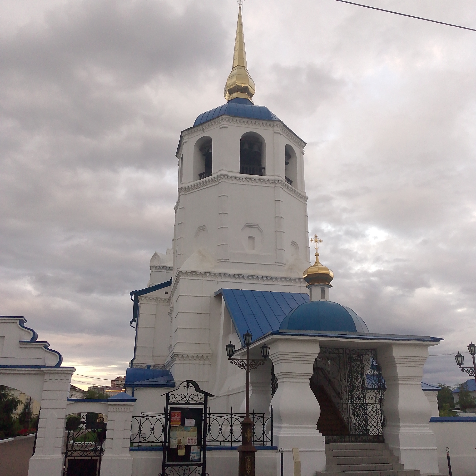 The image shows the bell tower of the Odigitrievsky Cathedral in Ulan-Ude, Russia. The bell tower is white with blue domes and a golden spire. The sky is overcast with gray clouds. There is a wrought-iron fence in front of the bell tower, and a street lamp on the right side of the image. In the foreground, there is a white arched gateway with blue trim. In the distance, there are some buildings visible.
